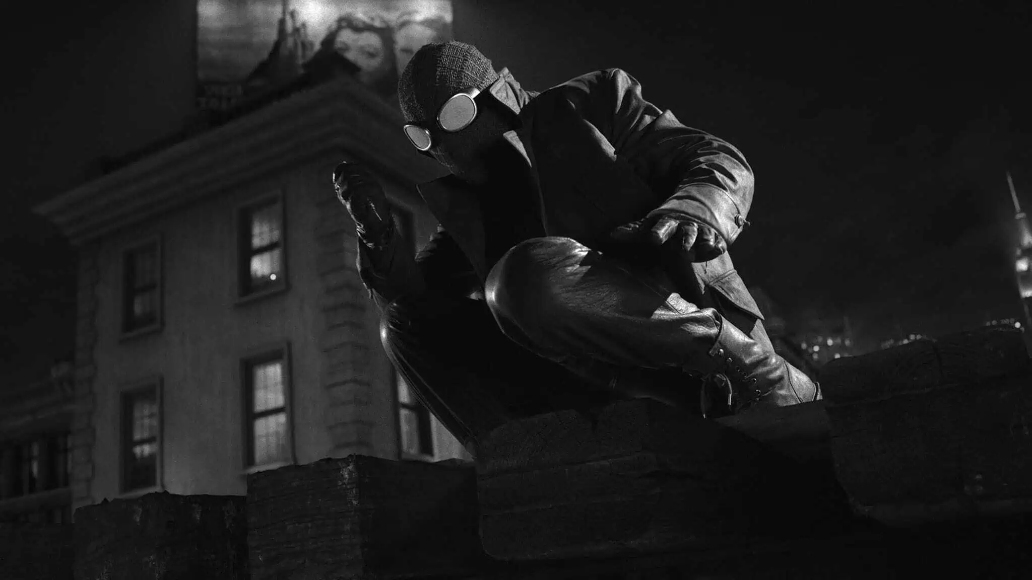 A person in a dark, rugged Spider-Man suit crouches on a rooftop at night in a black-and-white scene, with a building and a billboard visible in the background.