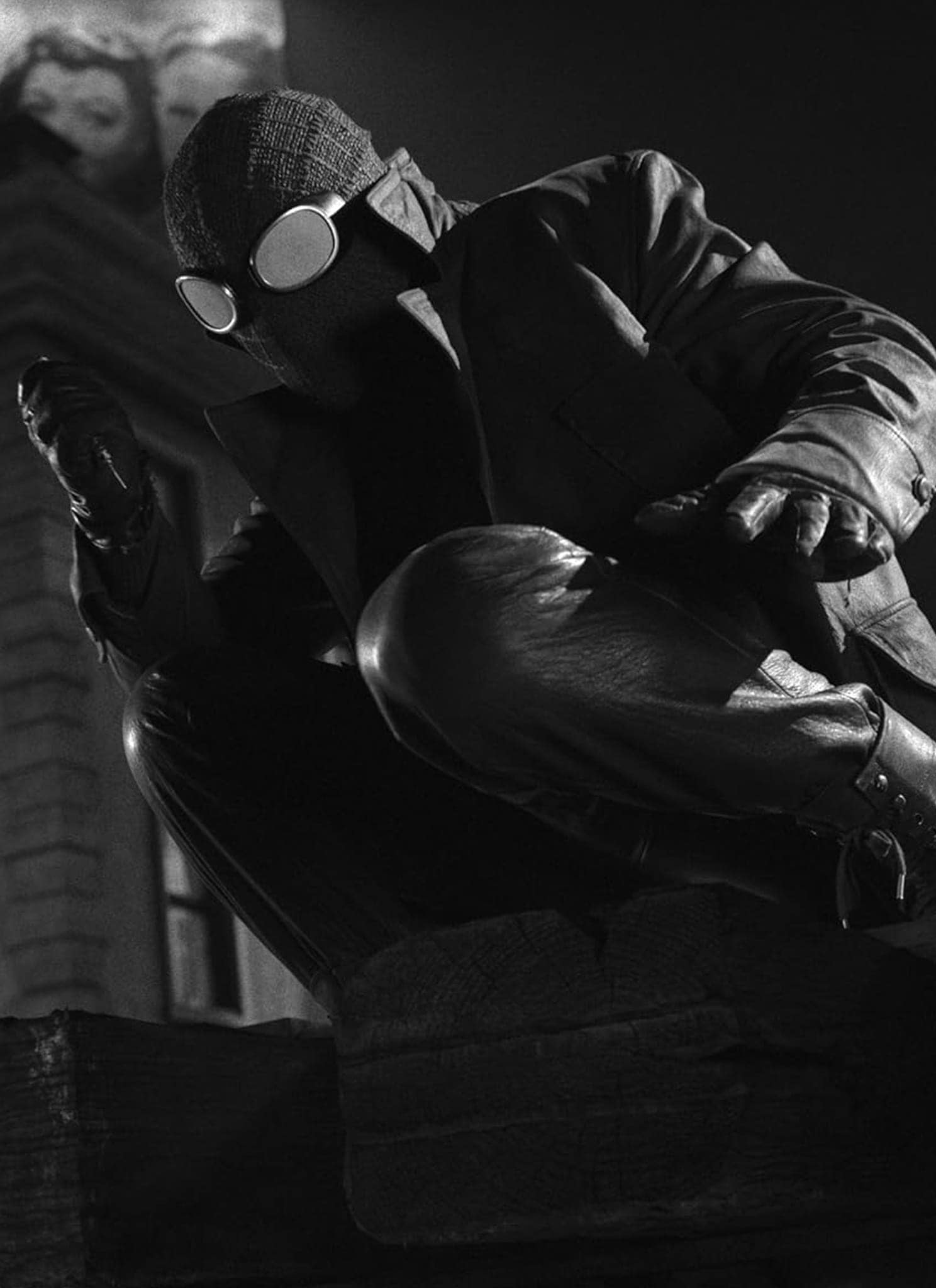A person in a dark, rugged Spider-Man suit crouches on a rooftop at night in a black-and-white scene, with a building and a billboard visible in the background.