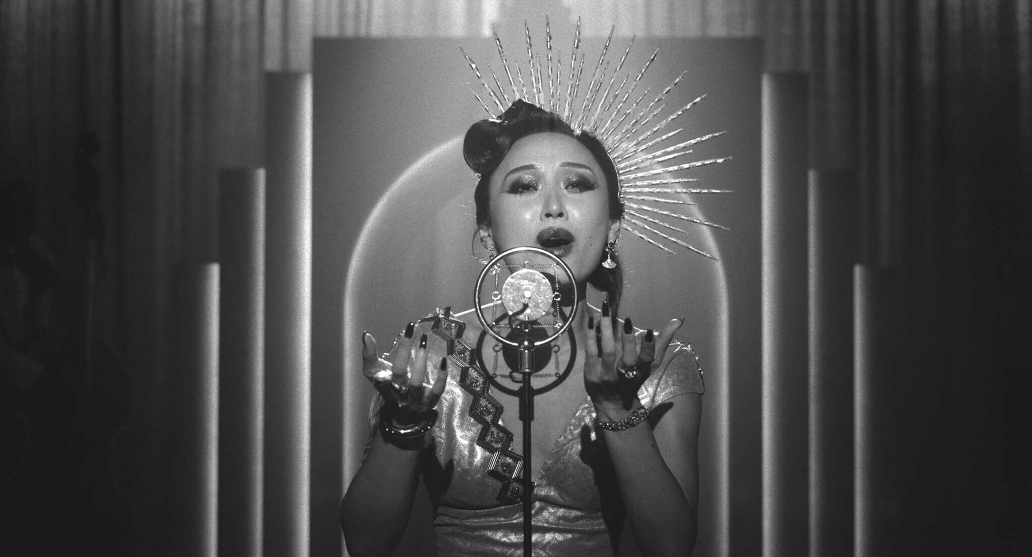 A woman in a vintage dress and spiked headpiece sings passionately into an old-fashioned microphone on a stage, lit by dramatic lighting in a black-and-white setting.