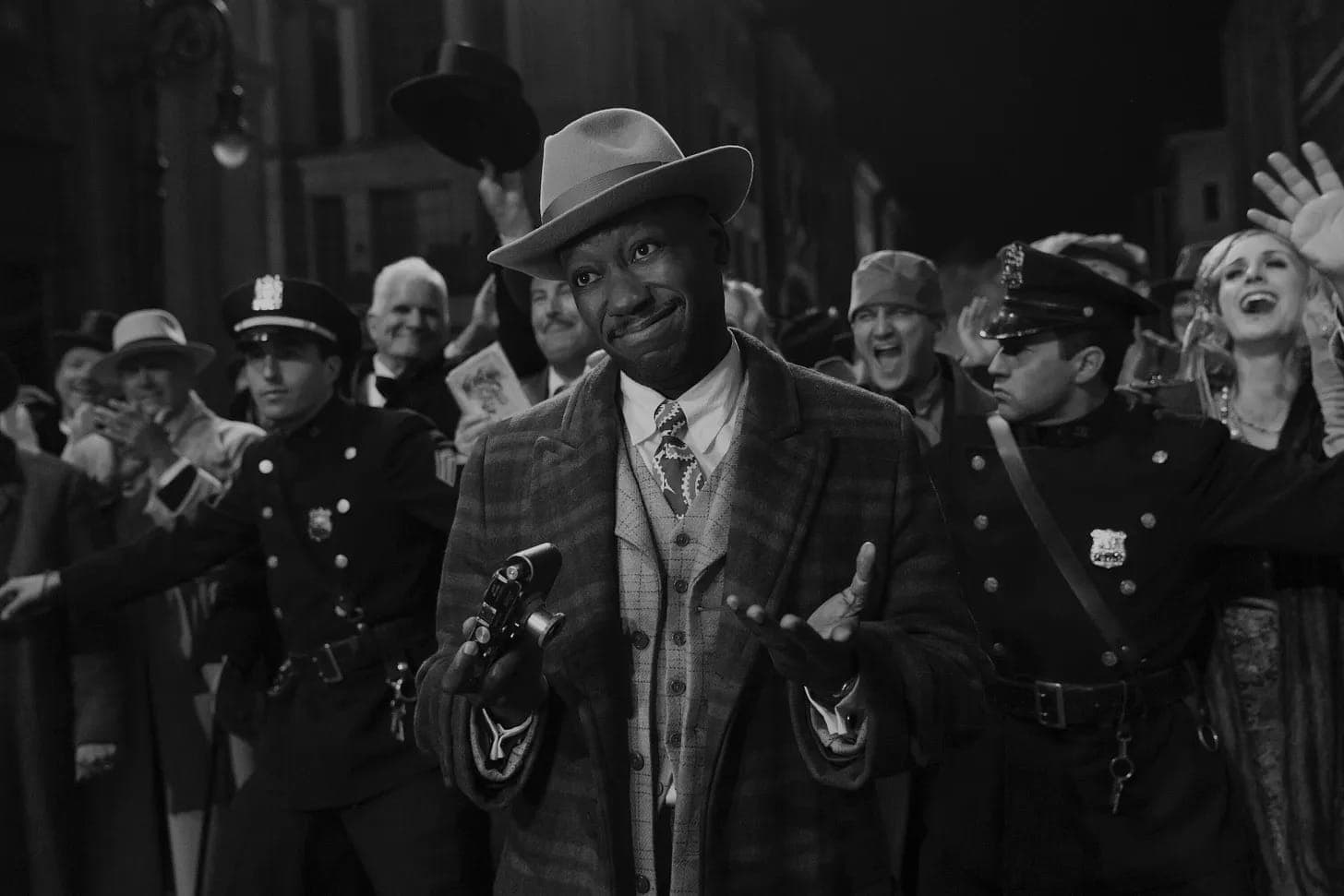 A man in vintage clothing shrugs and holds a camera while surrounded by cheering people and police officers in a crowded street at night; the image is in black and white.