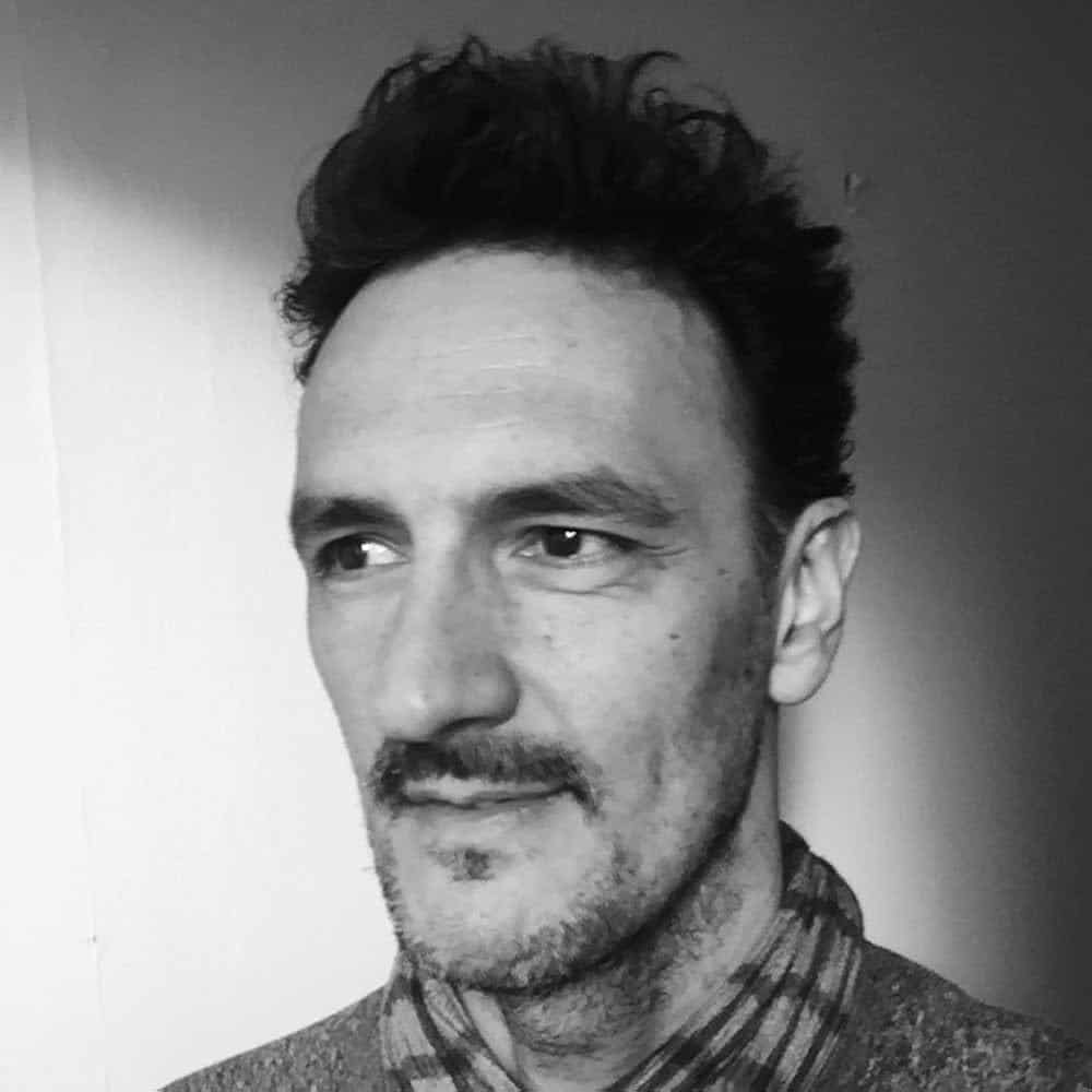 Black and white photo of a man with short, wavy hair and a light beard, wearing a scarf and sweater, looking slightly to the right against a plain wall background.
