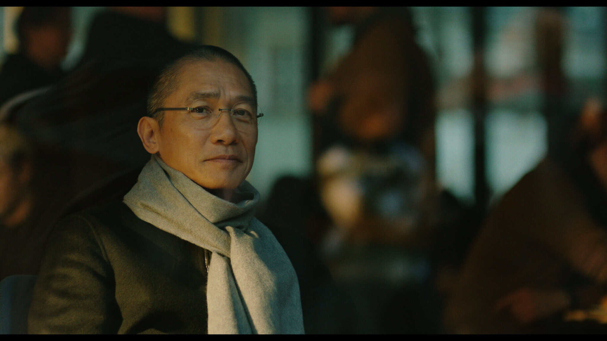 A man wearing glasses, a dark coat, and a light scarf sits indoors, looking calmly at the camera. The background is blurred with people and warm lighting.