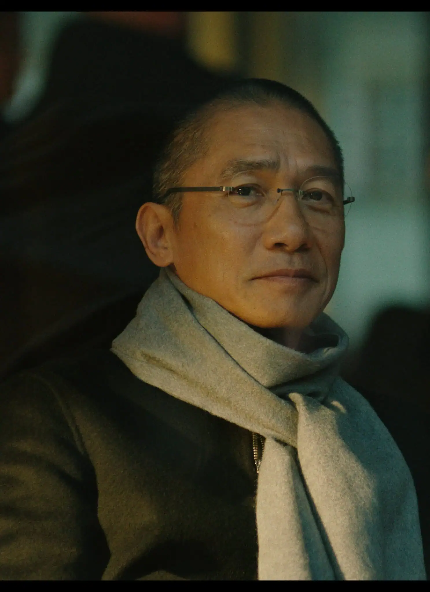 A man wearing glasses, a dark coat, and a light scarf sits indoors, looking calmly at the camera. The background is blurred with people and warm lighting.