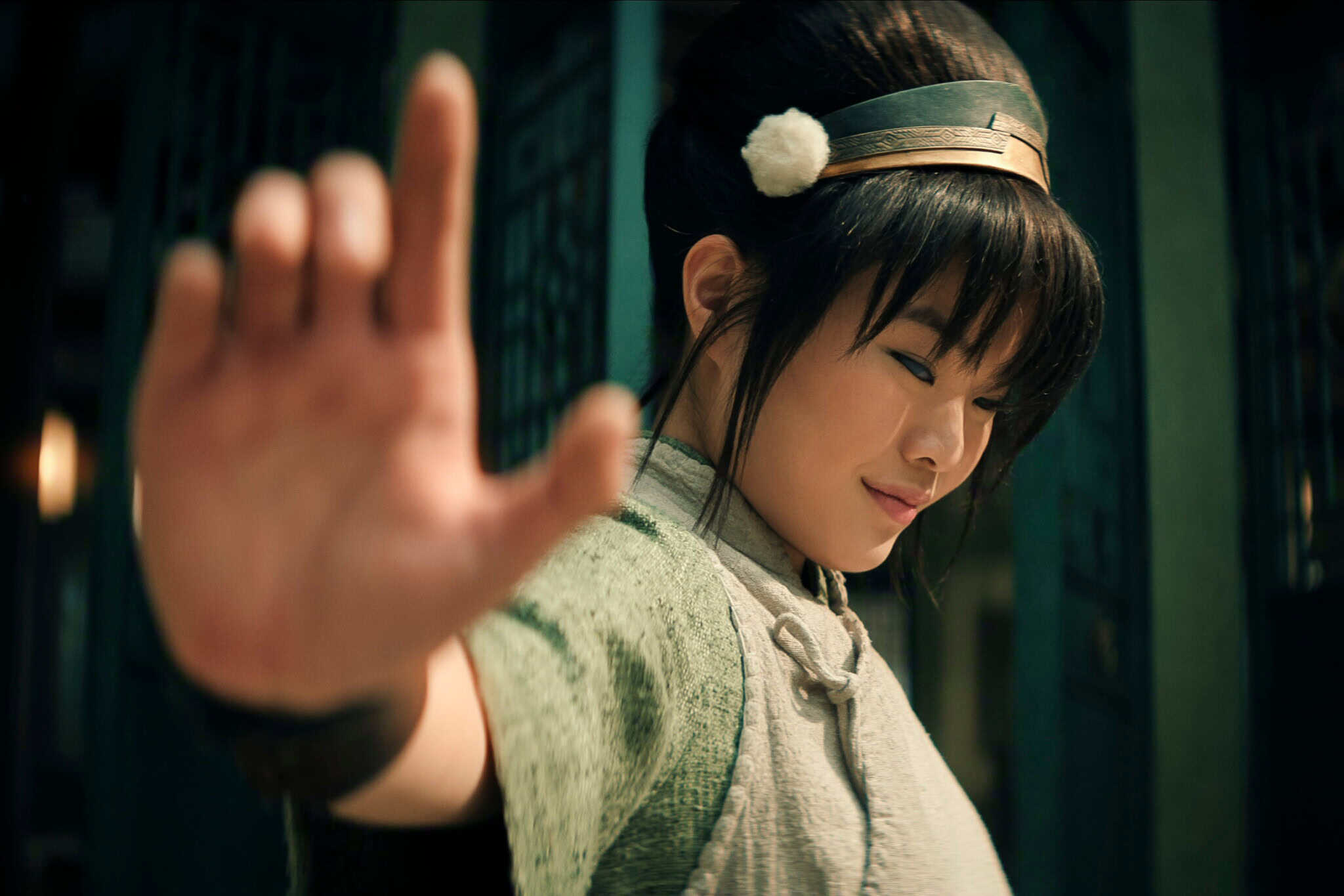 A young woman in traditional Asian clothing raises her hand in front of her, palm open, with a calm and focused expression, standing indoors against a blurred background.