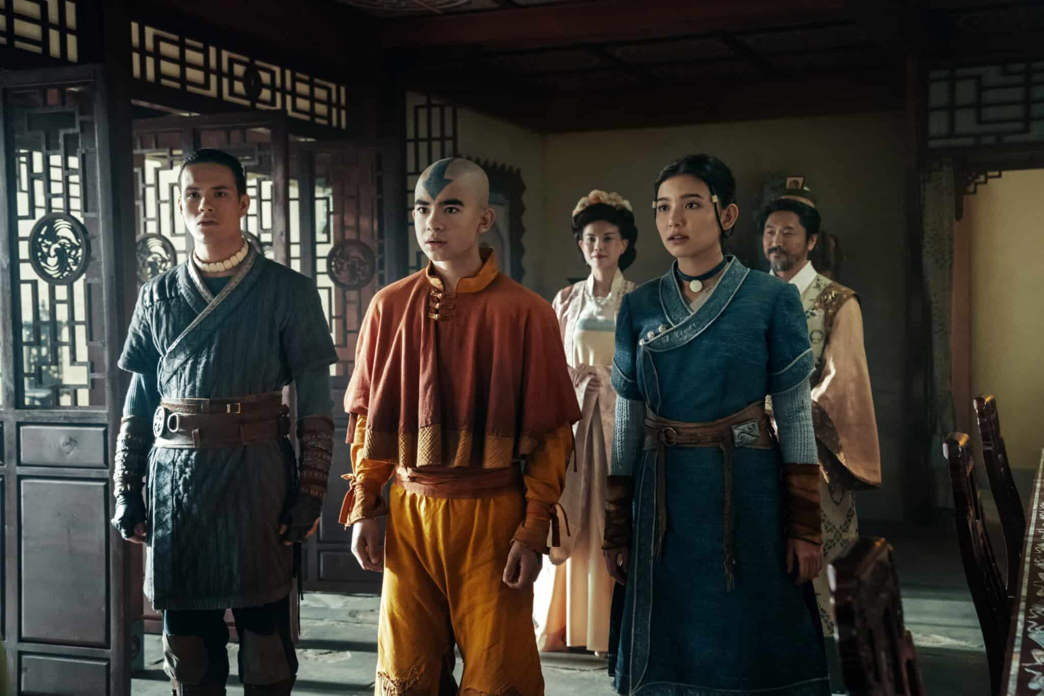 Three young people in detailed costumes stand side by side in a traditional, ornate room, with two adults in the background. All have serious expressions, and the central figure has a blue arrow tattoo on his head.