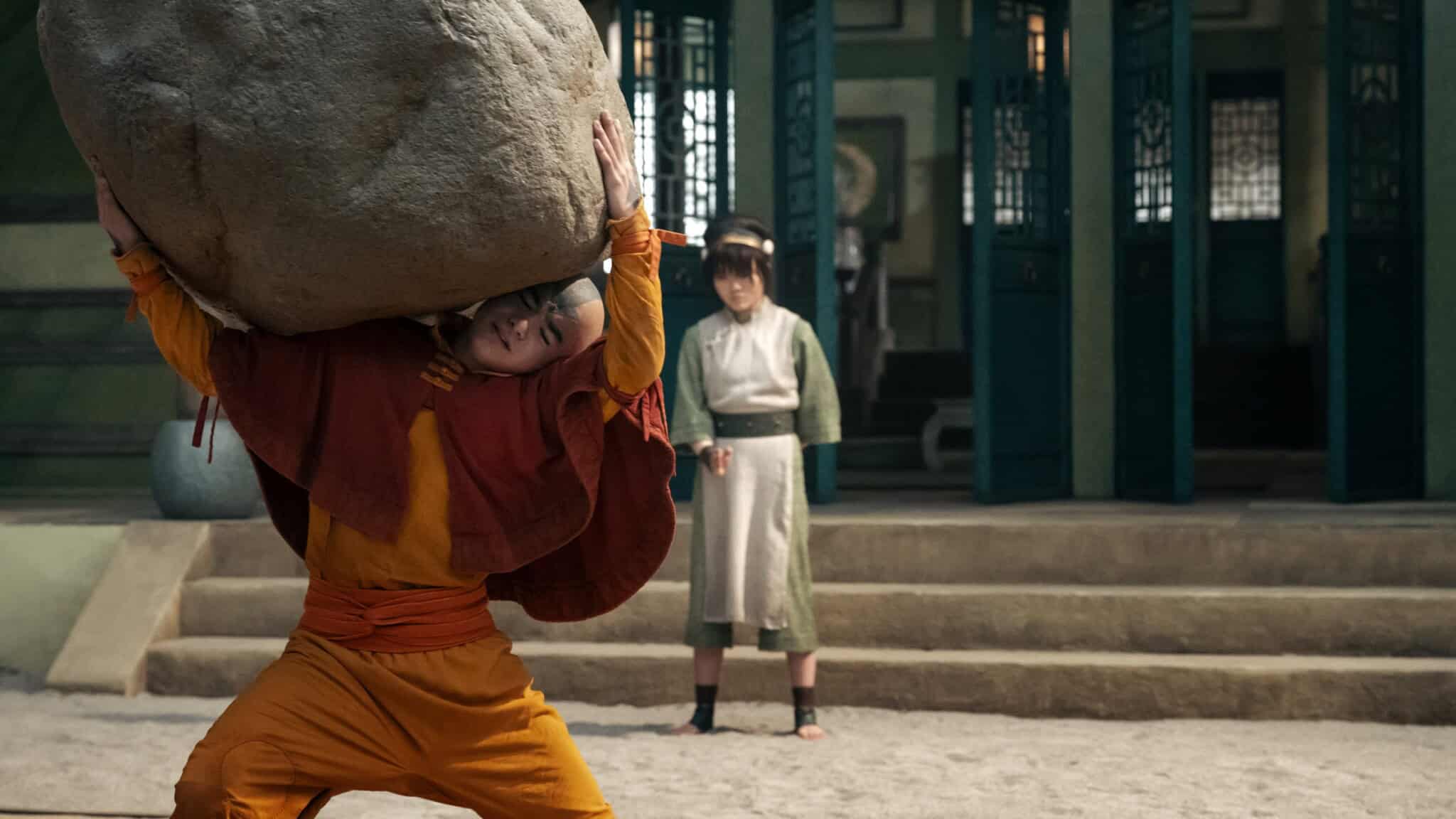 A person dressed in orange and yellow lifts a large boulder above their head, while another person in green and white watches from nearby steps in a courtyard.