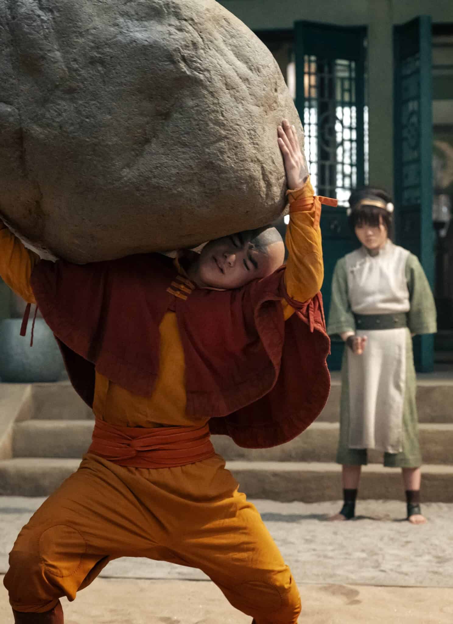 A person dressed in orange and yellow lifts a large boulder above their head, while another person in green and white watches from nearby steps in a courtyard.