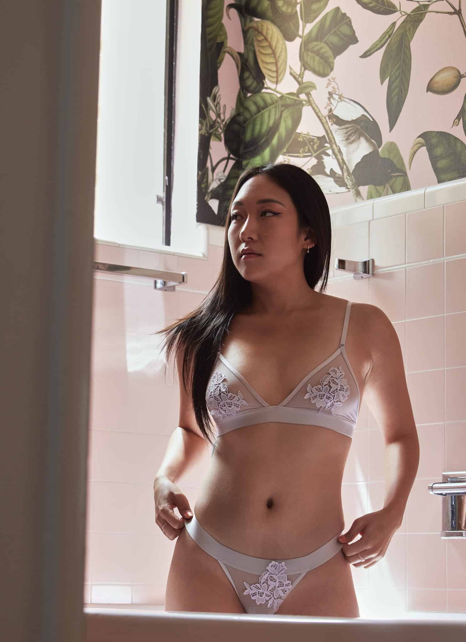 A woman with long dark hair wearing a light gray bra with floral lace stands in a tiled bathroom, looking thoughtfully to the side with soft sunlight coming through a window.