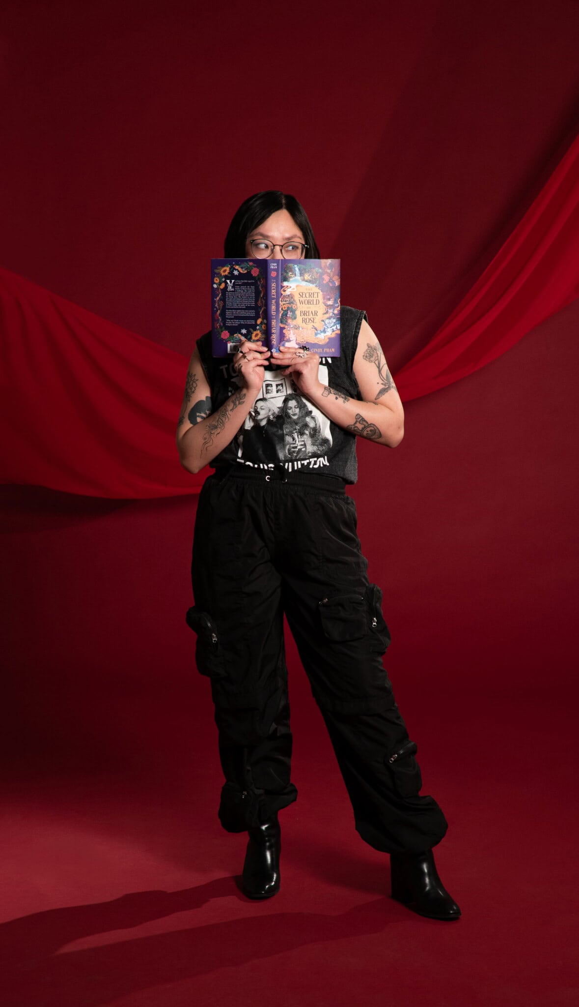 A person with black hair and glasses stands against a red backdrop, holding an open book in front of their face. They wear a black graphic t-shirt, cargo pants, and boots, showing several tattoos on their arms.
