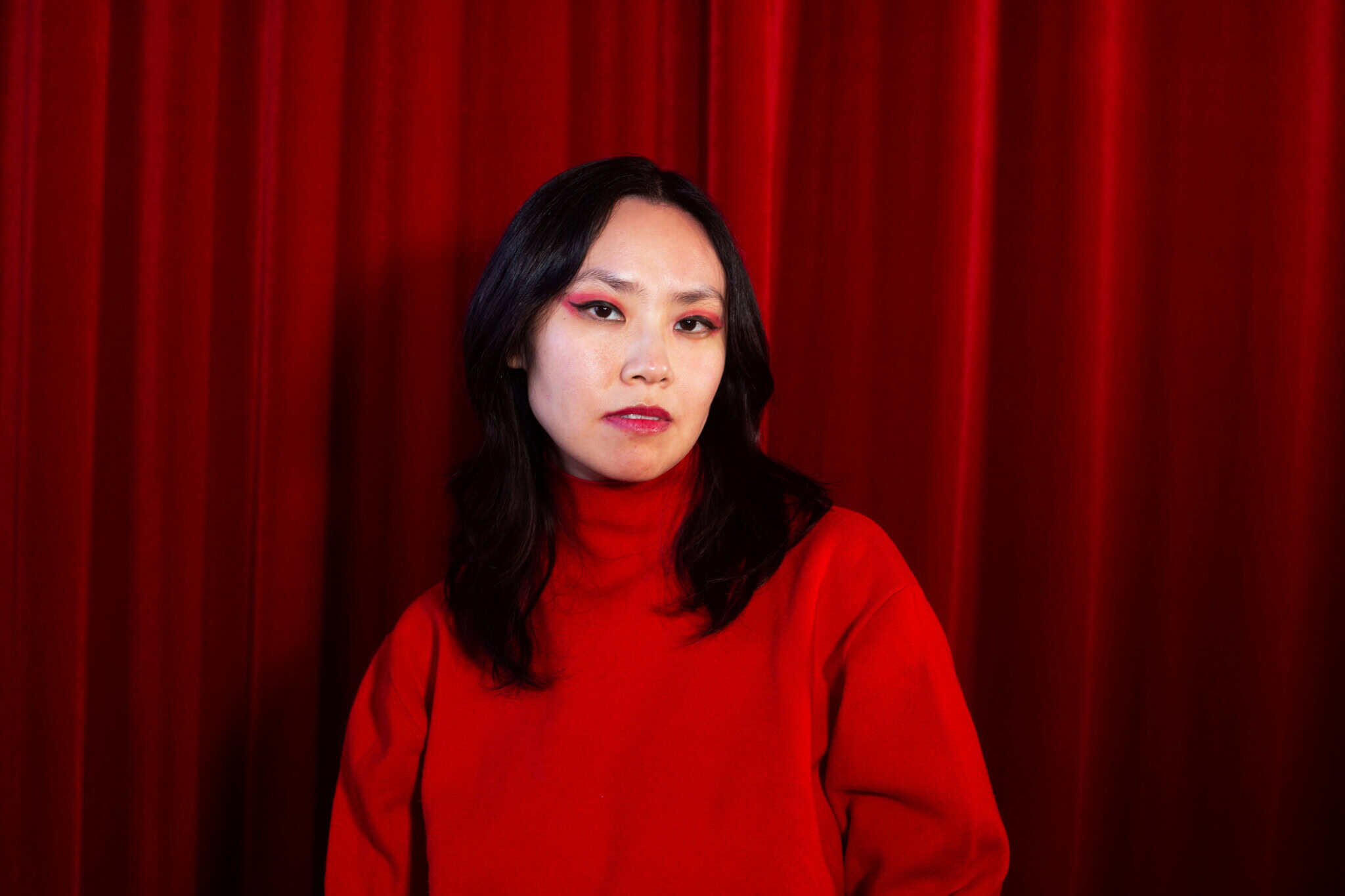 A person with long dark hair, wearing a red turtleneck sweater and bold red eye makeup, stands in front of red curtains, looking directly at the camera with a neutral expression.
