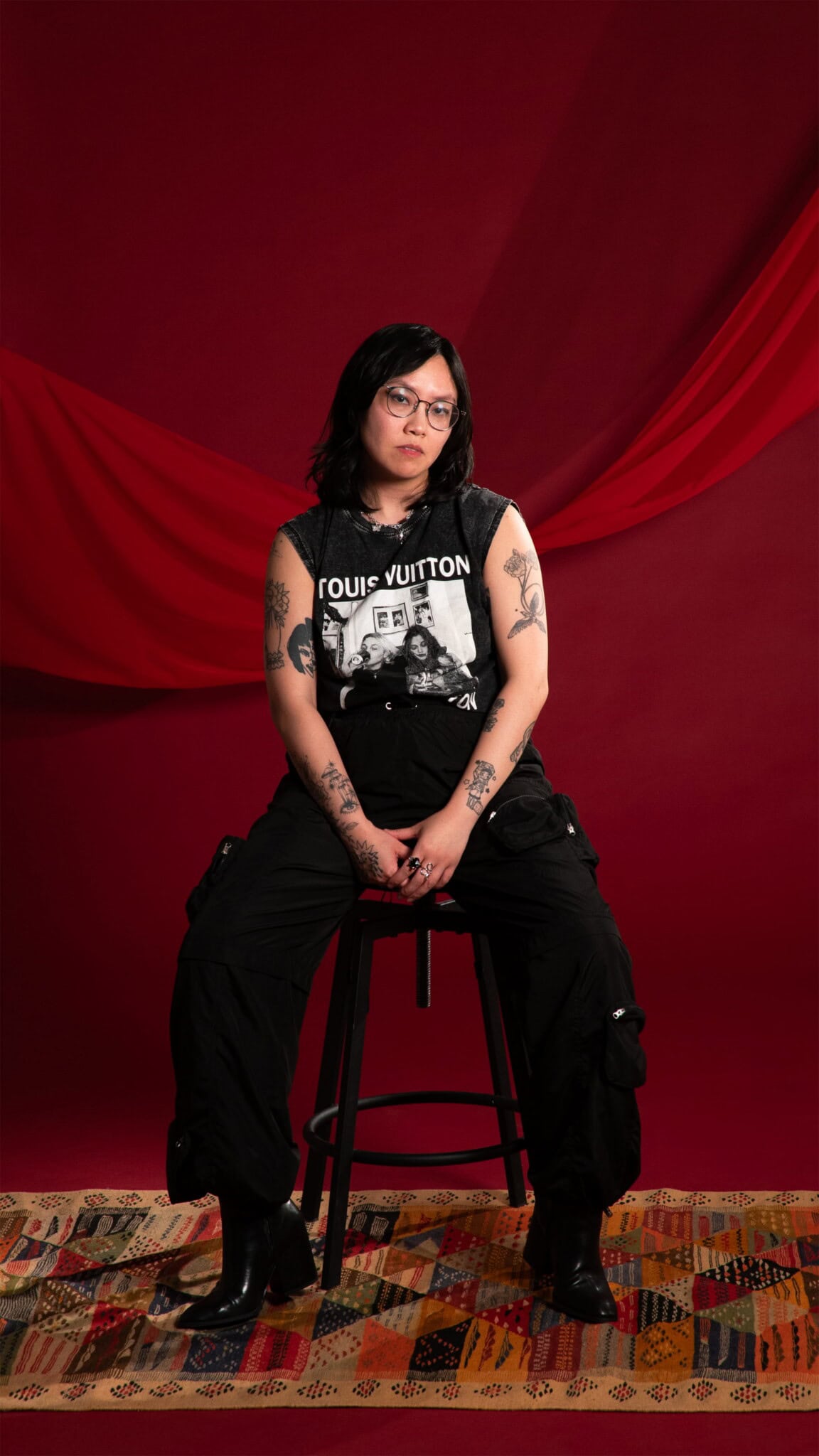 A person with glasses and tattoos sits on a stool against a red backdrop with draped fabric, wearing a black sleeveless shirt, black cargo pants, and black boots on a colorful patterned rug.