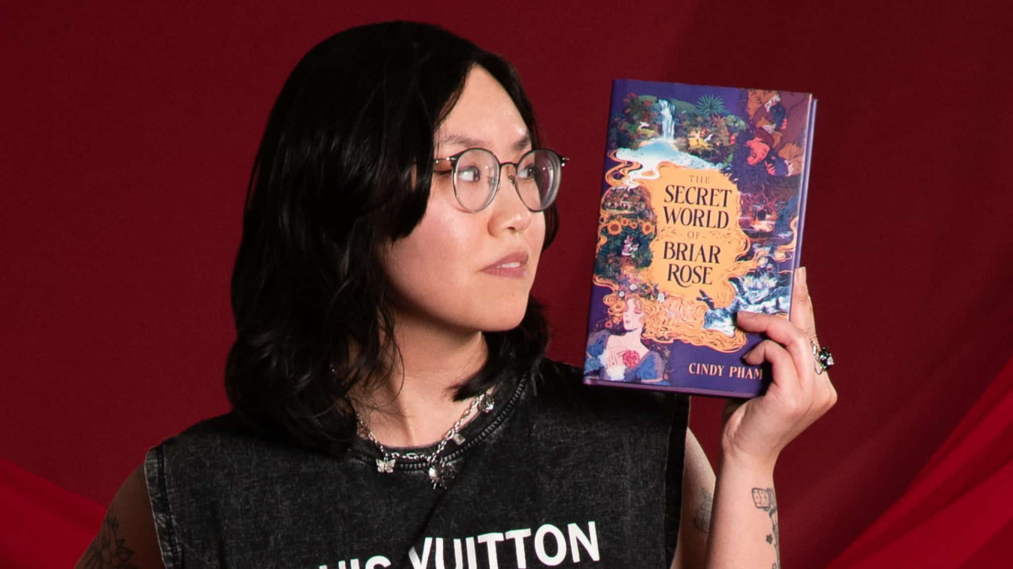 A person with glasses and long dark hair holds up a colorful book titled The Secret World of Briar Rose by Cindy Phan, against a red background. They wear a black sleeveless shirt and layered necklaces.