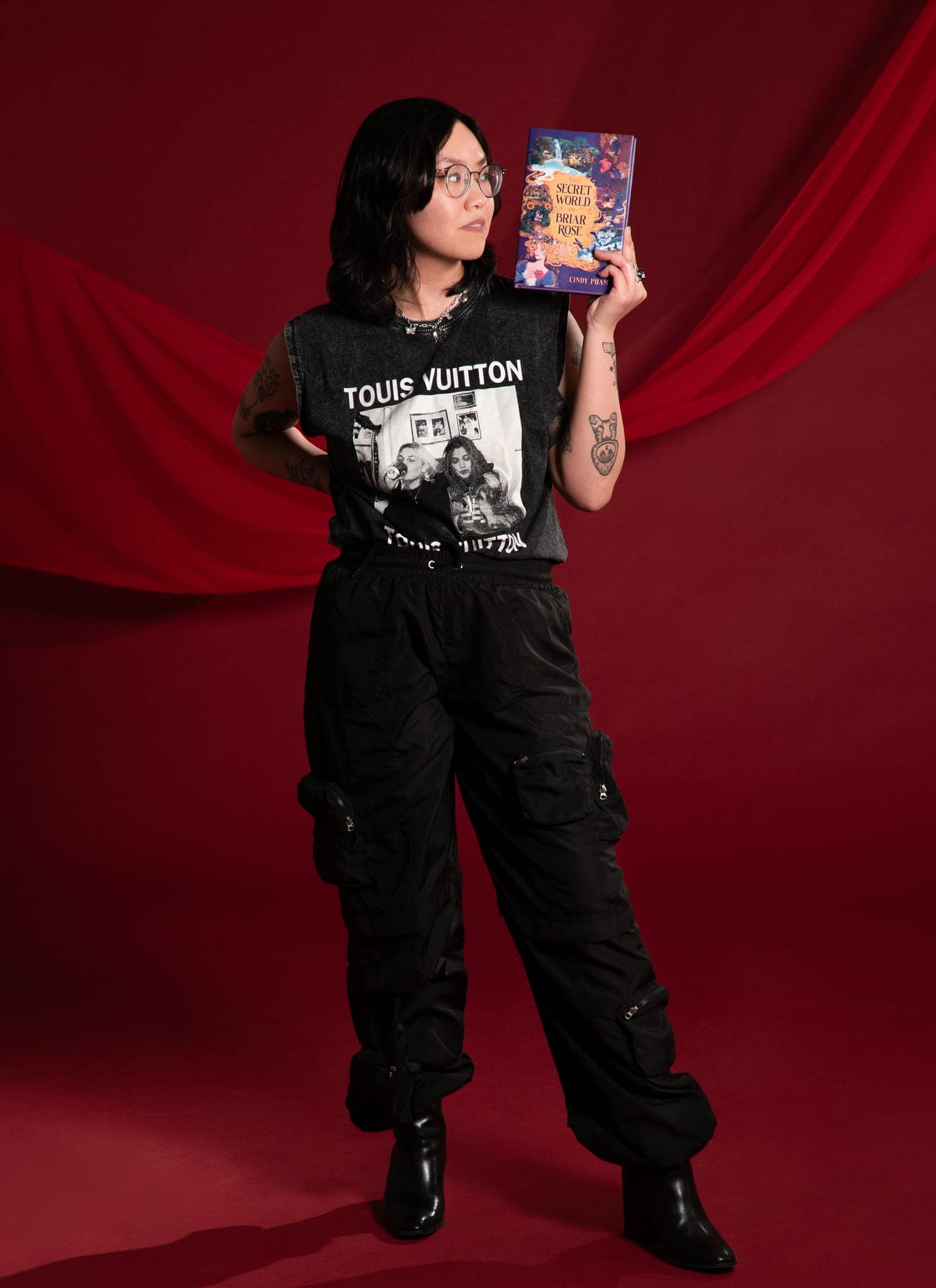 A person with glasses and long dark hair holds up a colorful book titled The Secret World of Briar Rose by Cindy Phan, against a red background. They wear a black sleeveless shirt and layered necklaces.