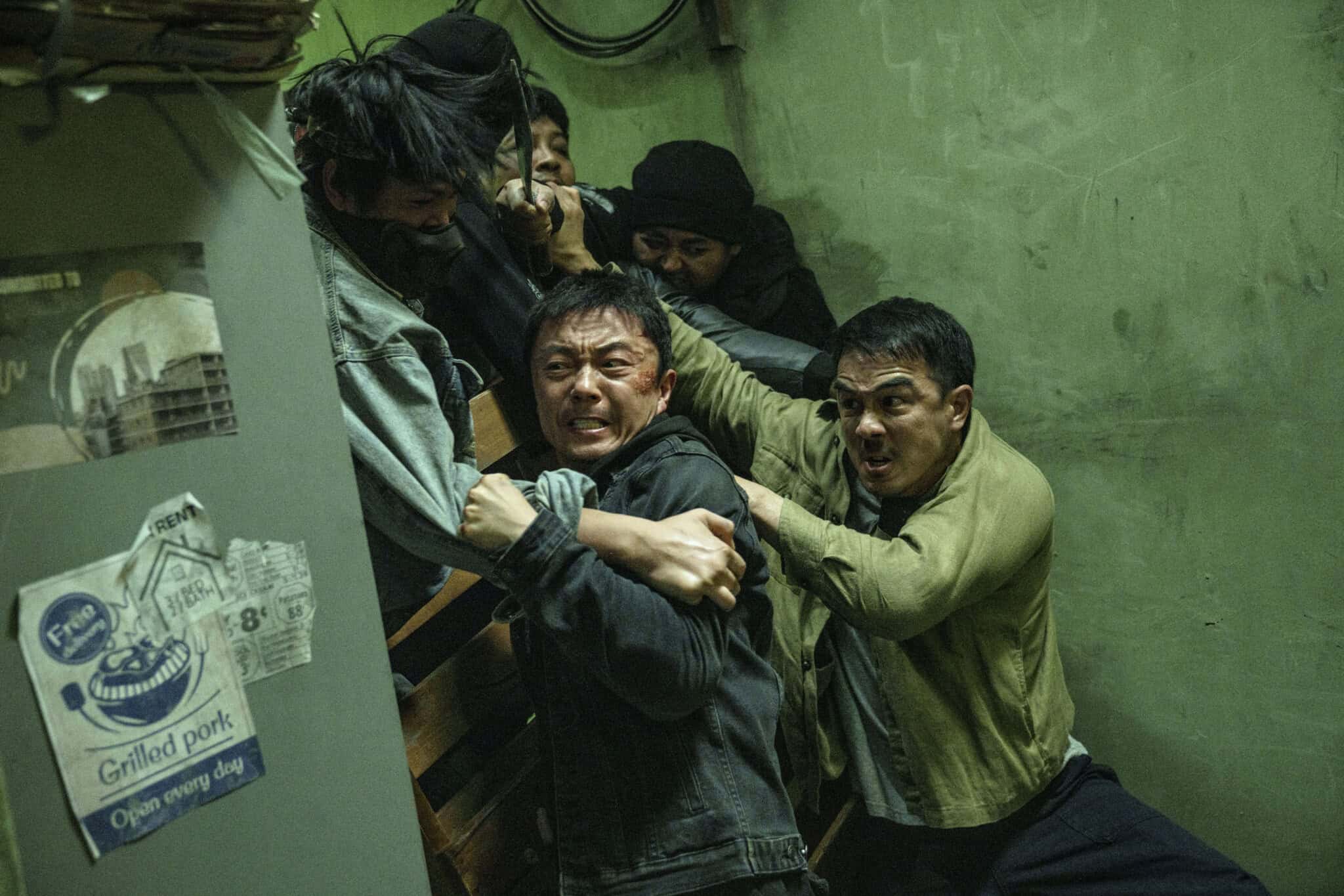 Several men are engaged in an intense struggle in a dimly lit, green-walled room, their faces showing strain as they grapple and push each other near a cluttered corner with a Grilled pork poster on the wall.
