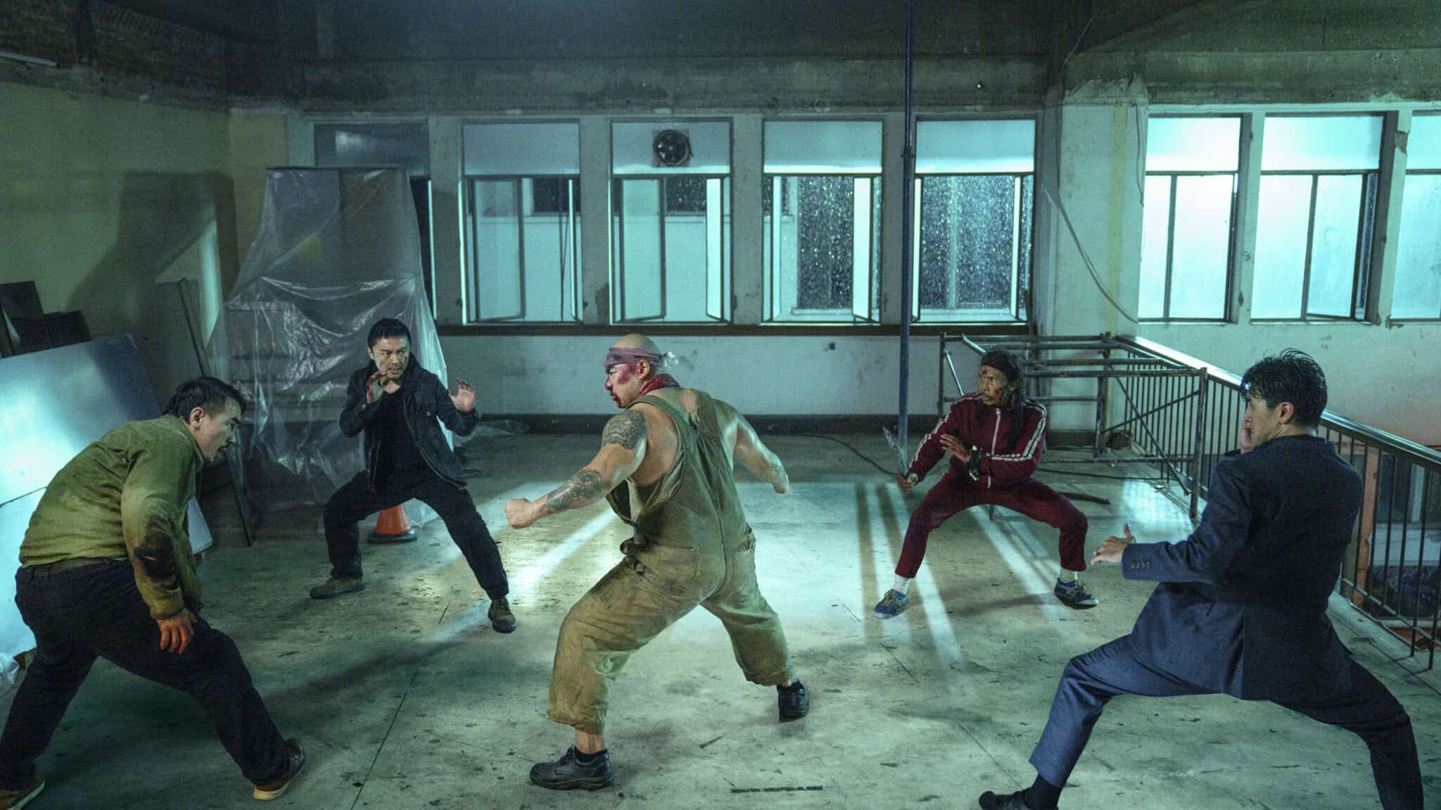 Five men in a dimly lit, industrial room stand in fighting stances, facing each other as if preparing for a fight scene. The atmosphere is tense, with scattered debris and blue-tinted lighting.