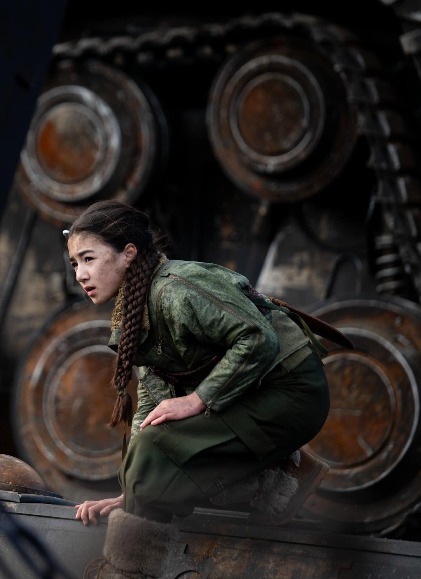 A young woman with braided hair, dressed in green, crouches on a large industrial machine with massive gears and treads in the background, her focused gaze reminiscent of Supergirl's unwavering determination.