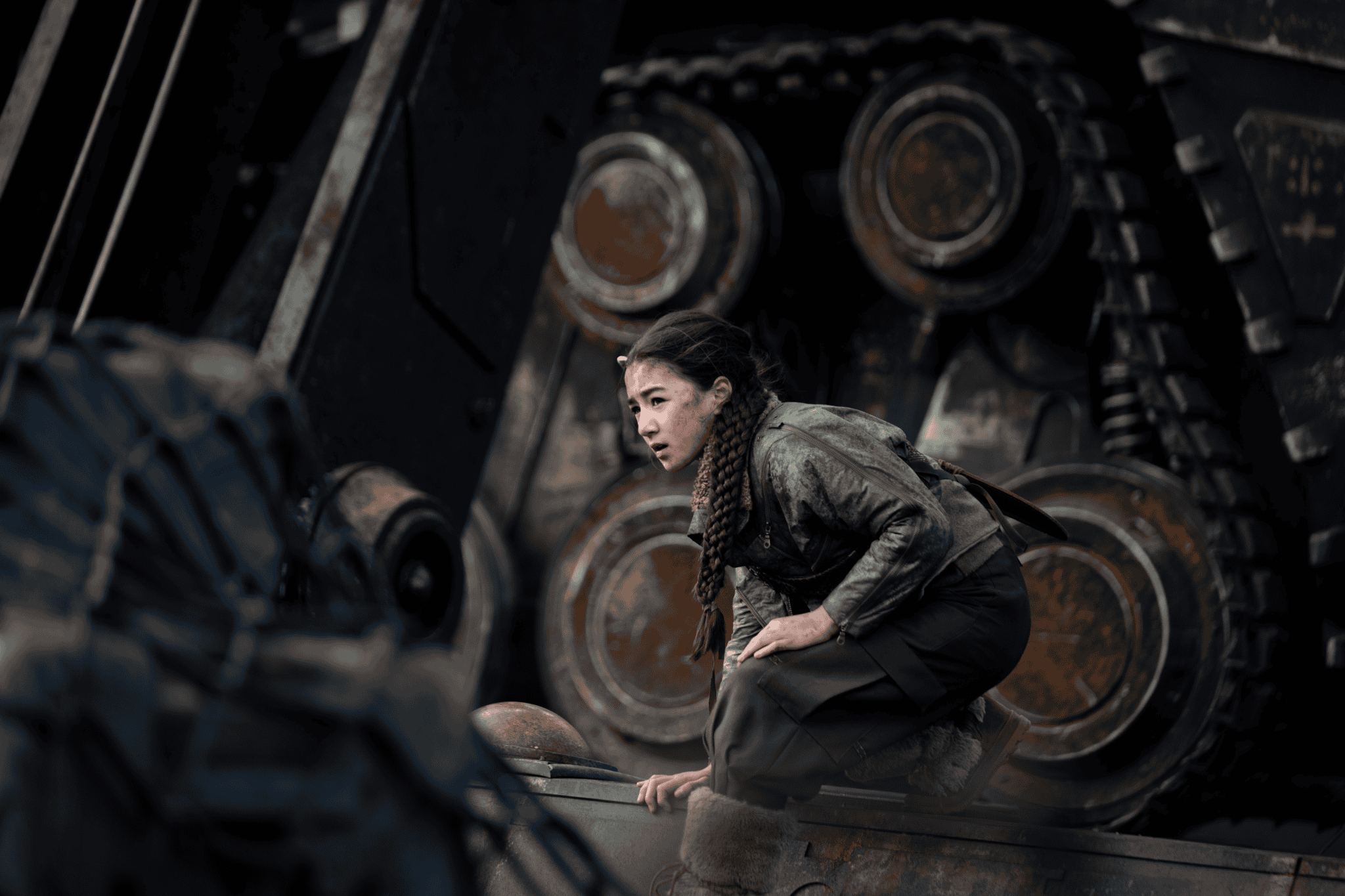A young woman with braided hair, dressed in green, crouches on a large industrial machine with massive gears and treads in the background, her focused gaze reminiscent of Supergirl's unwavering determination.