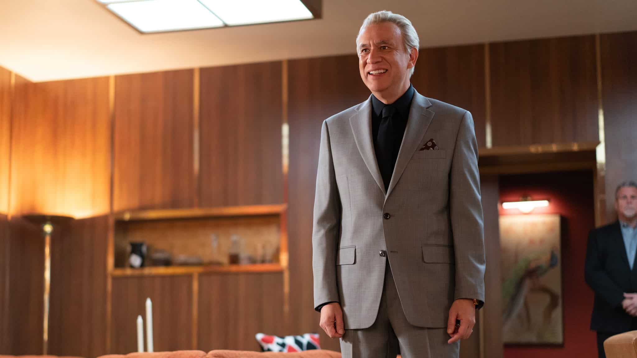 A man in a light gray suit stands and smiles in a stylish, wood-paneled room with modern decor and warm lighting—a setting reminiscent of the classic charm embraced by the deli boys. Another man stands in the background near a red wall with a painting.