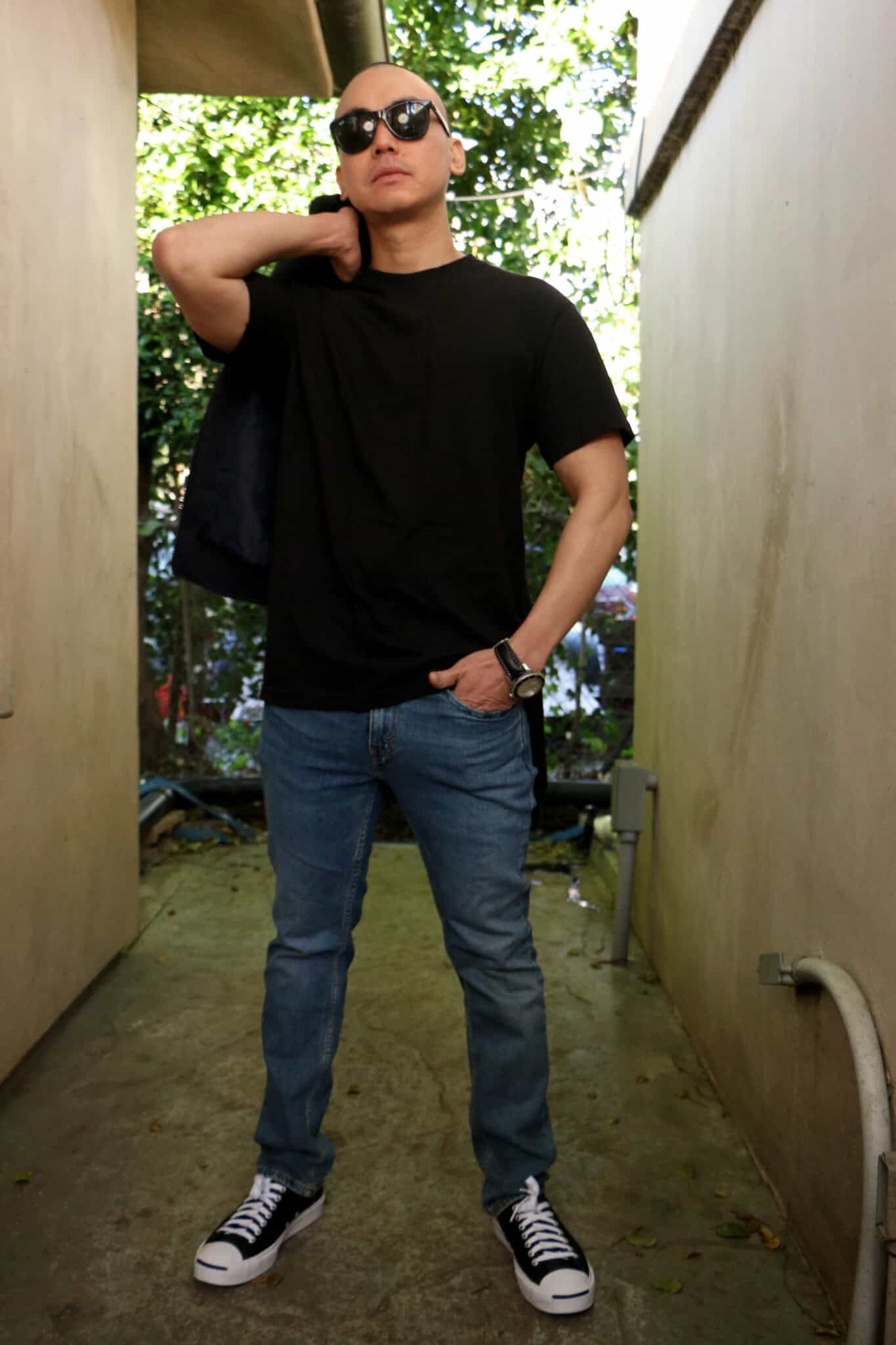 James Bang, wearing sunglasses, a black t-shirt, jeans, and black-and-white sneakers, stands confidently in a narrow outdoor corridor with one hand on his shoulder holding a jacket and the other in his pocket.