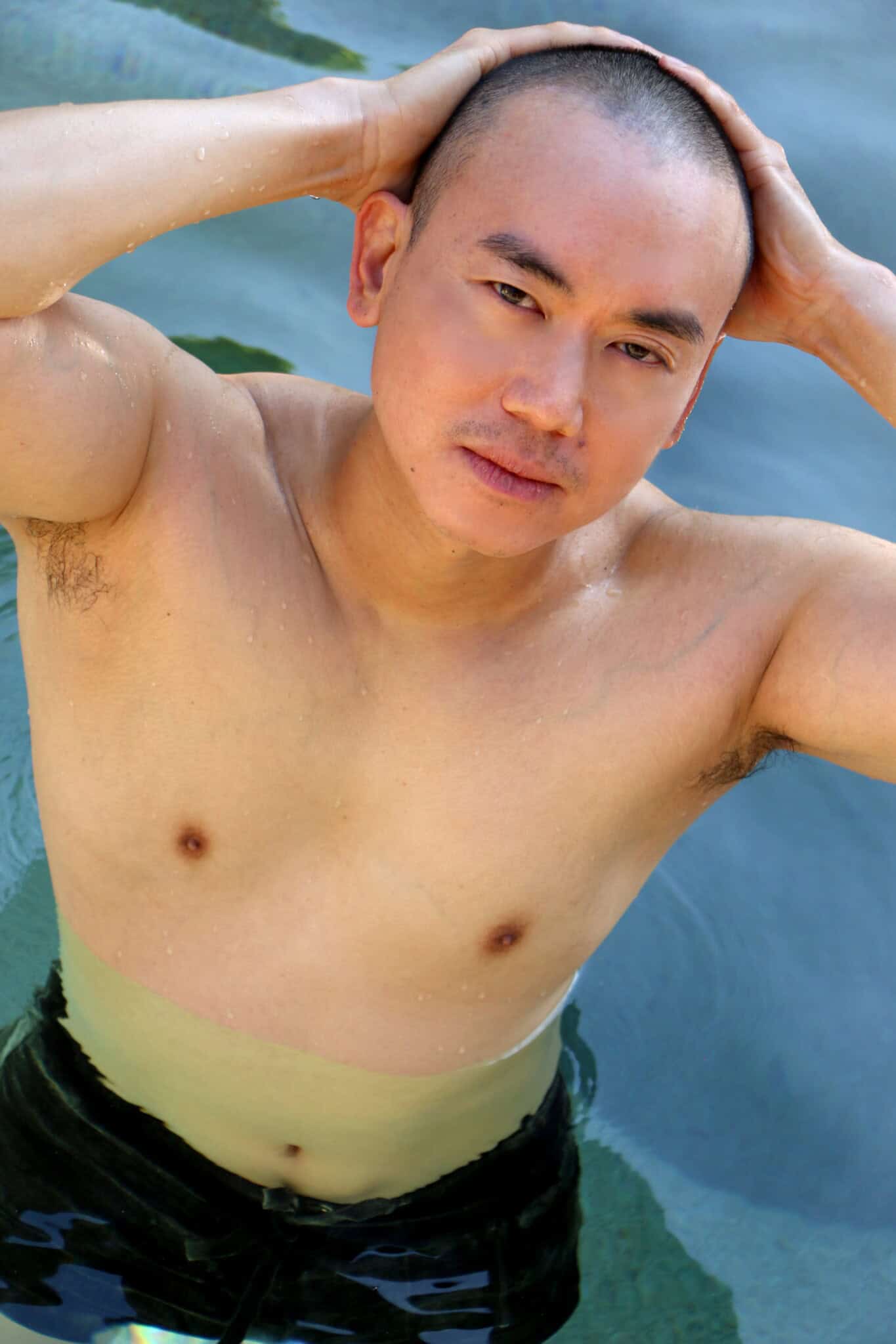 A shirtless person with short hair stands in water, wearing black shorts, with both hands resting on their head and looking up at the camera.
