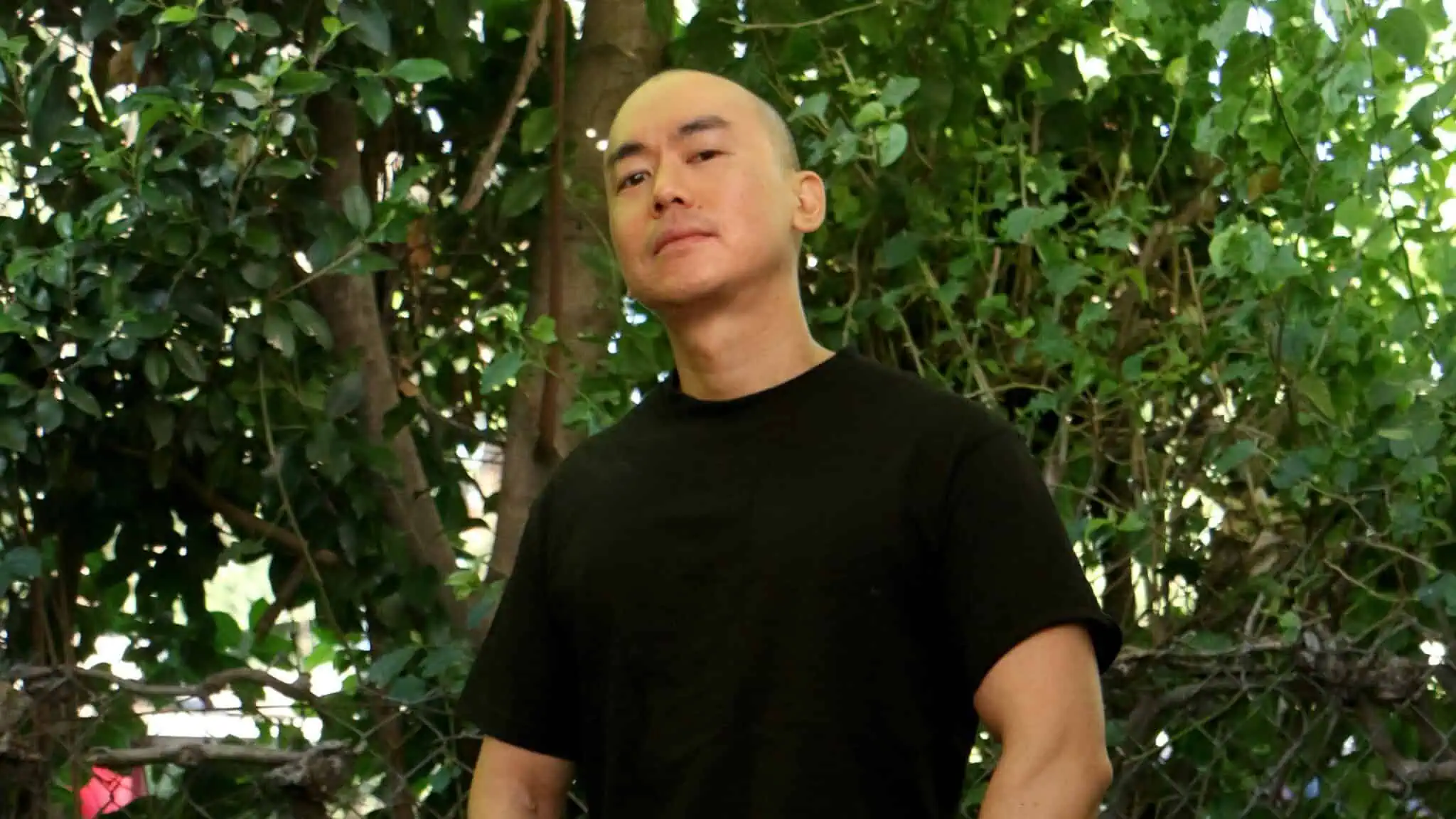 A person with a shaved head, wearing a black t-shirt, stands outside in front of dense green foliage, looking confidently at the camera.