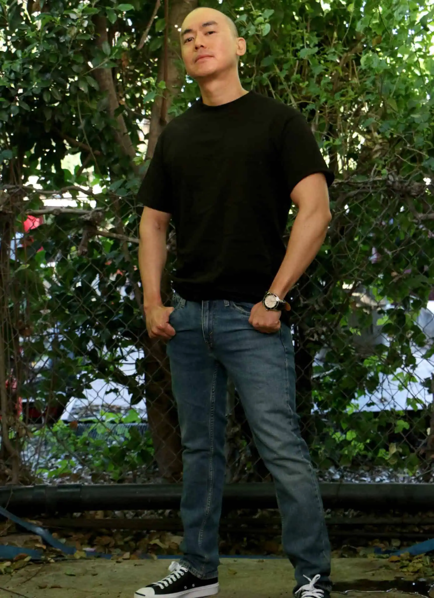 A person with a shaved head, wearing a black t-shirt, stands outside in front of dense green foliage, looking confidently at the camera.