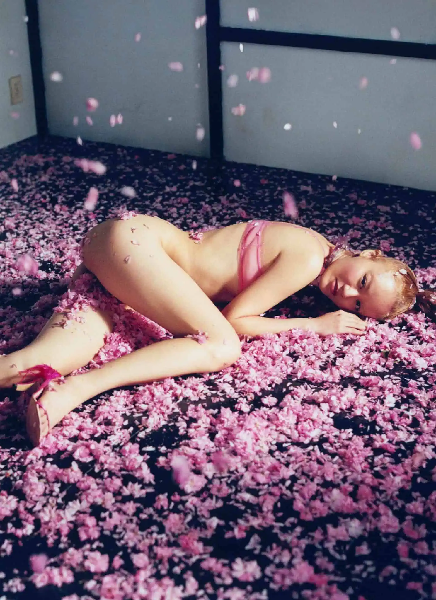 A woman wearing pink lingerie lies on her side on a dark floor covered in pink flower petals, with more petals falling around her in a softly lit room.