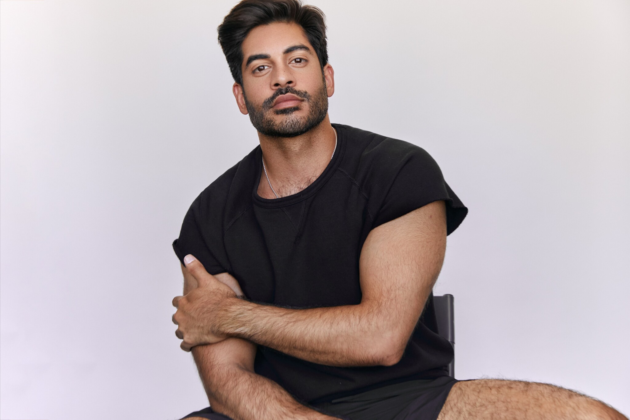 Sachin Bhatt, a man with short dark hair and a beard, sits on a chair against a plain background, wearing a black t-shirt and shorts, one arm resting across his body and a relaxed expression on his face.