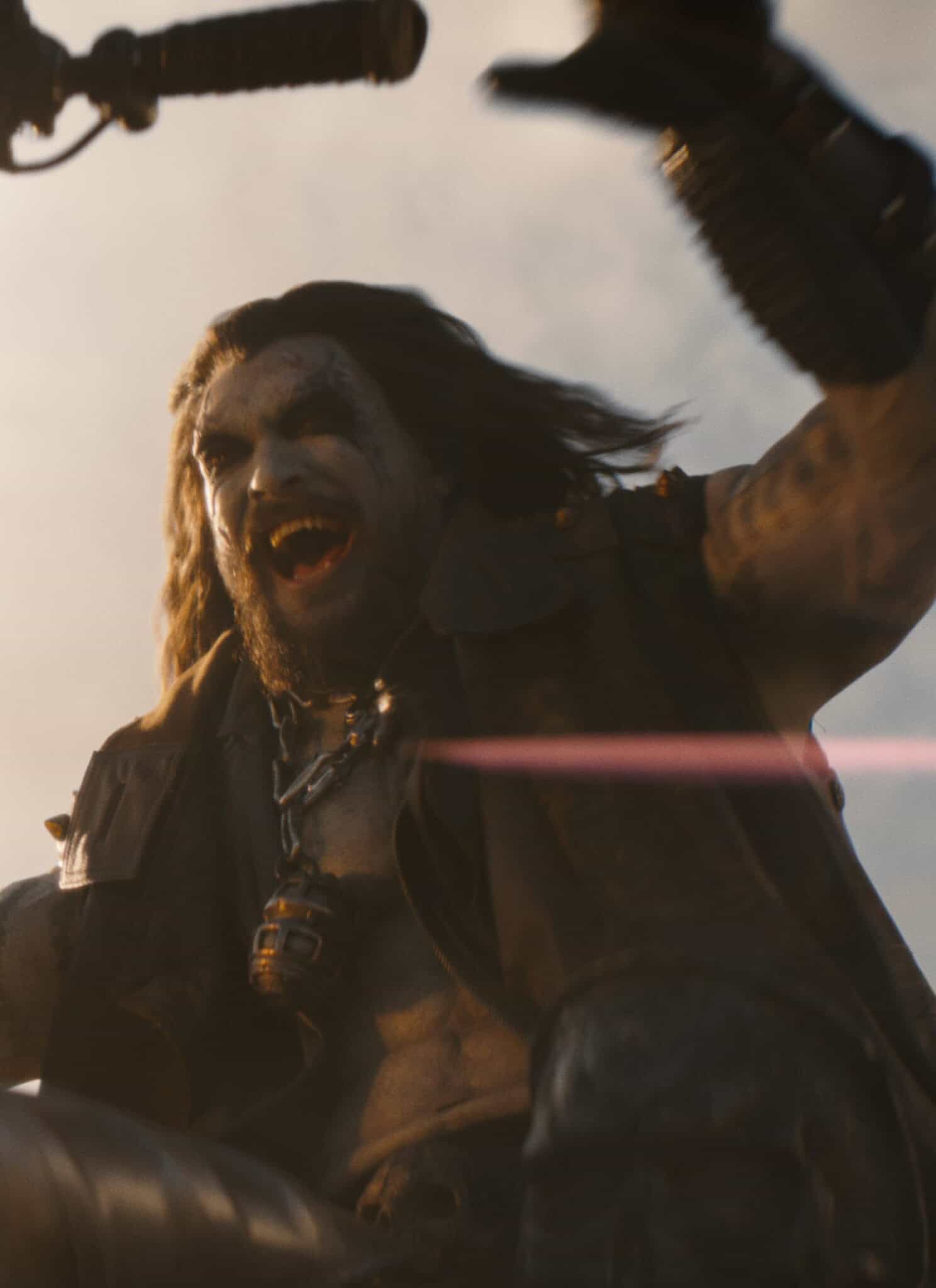 A rugged, fierce-looking man with long hair rides a motorcycle at high speed, yelling with excitement. The sun shines brightly behind him, and his arm is raised in the air.