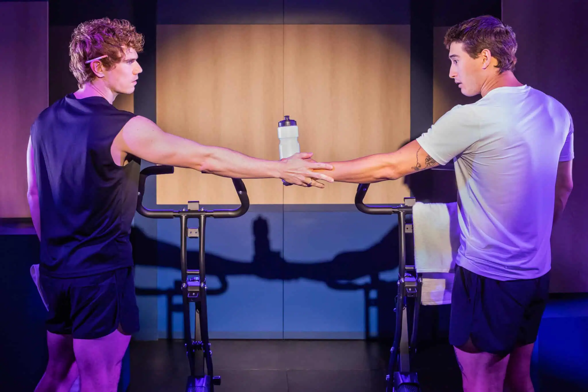Two men in athletic wear stand next to stationary bikes indoors, facing each other and passing a water bottle between them. One man wears black, the other white; a towel hangs on one bike.