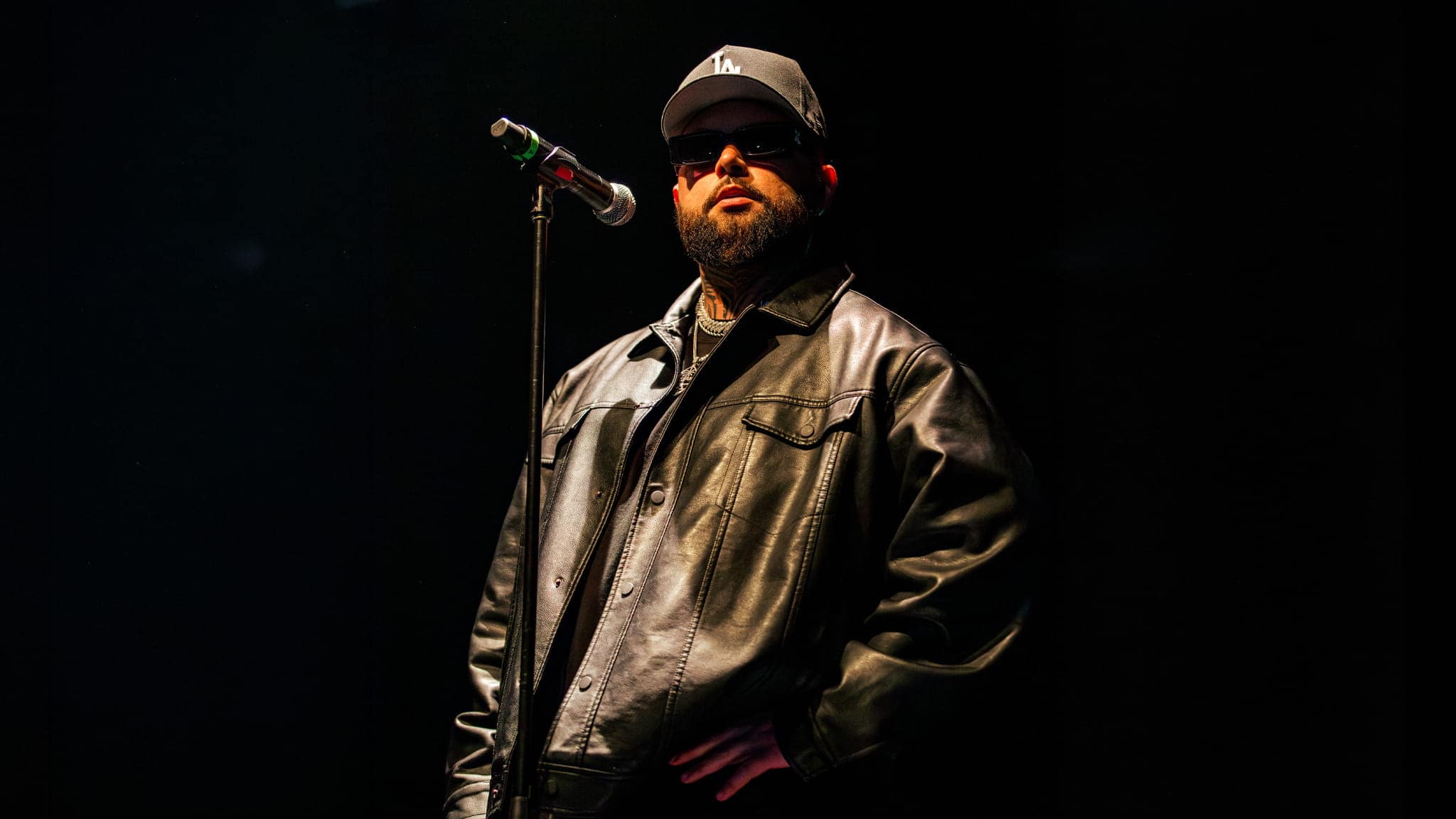 A man in a black leather jacket, black cap, and sunglasses stands on stage in front of a microphone, with one hand in his pocket against a dark background.