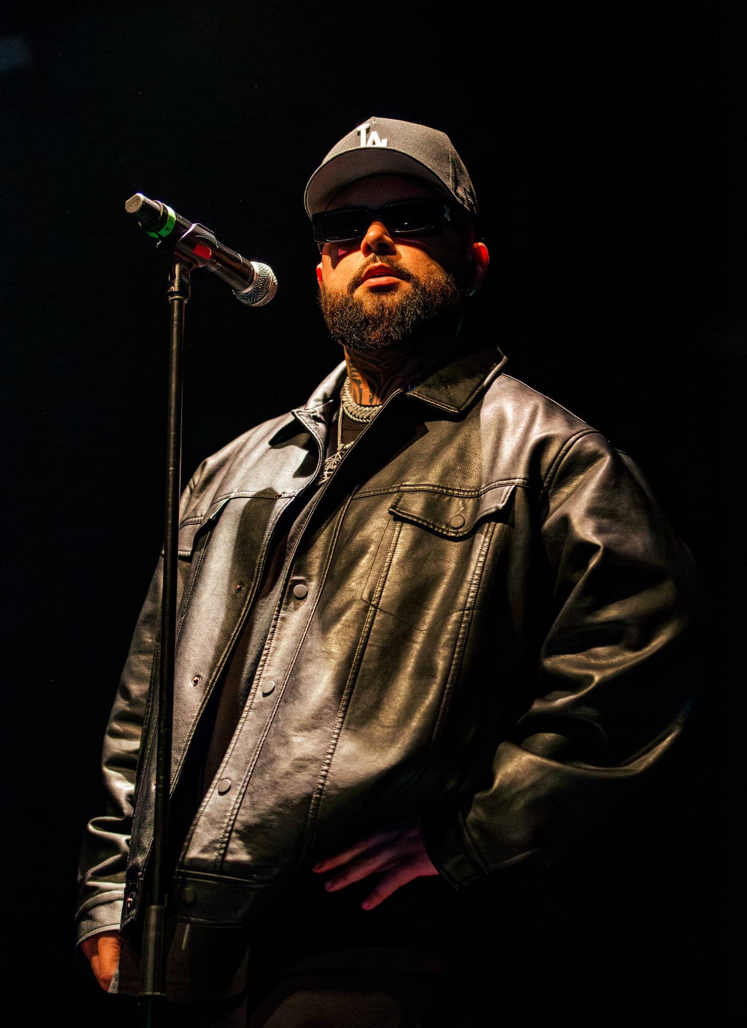 A man in a black leather jacket, black cap, and sunglasses stands on stage in front of a microphone, with one hand in his pocket against a dark background.
