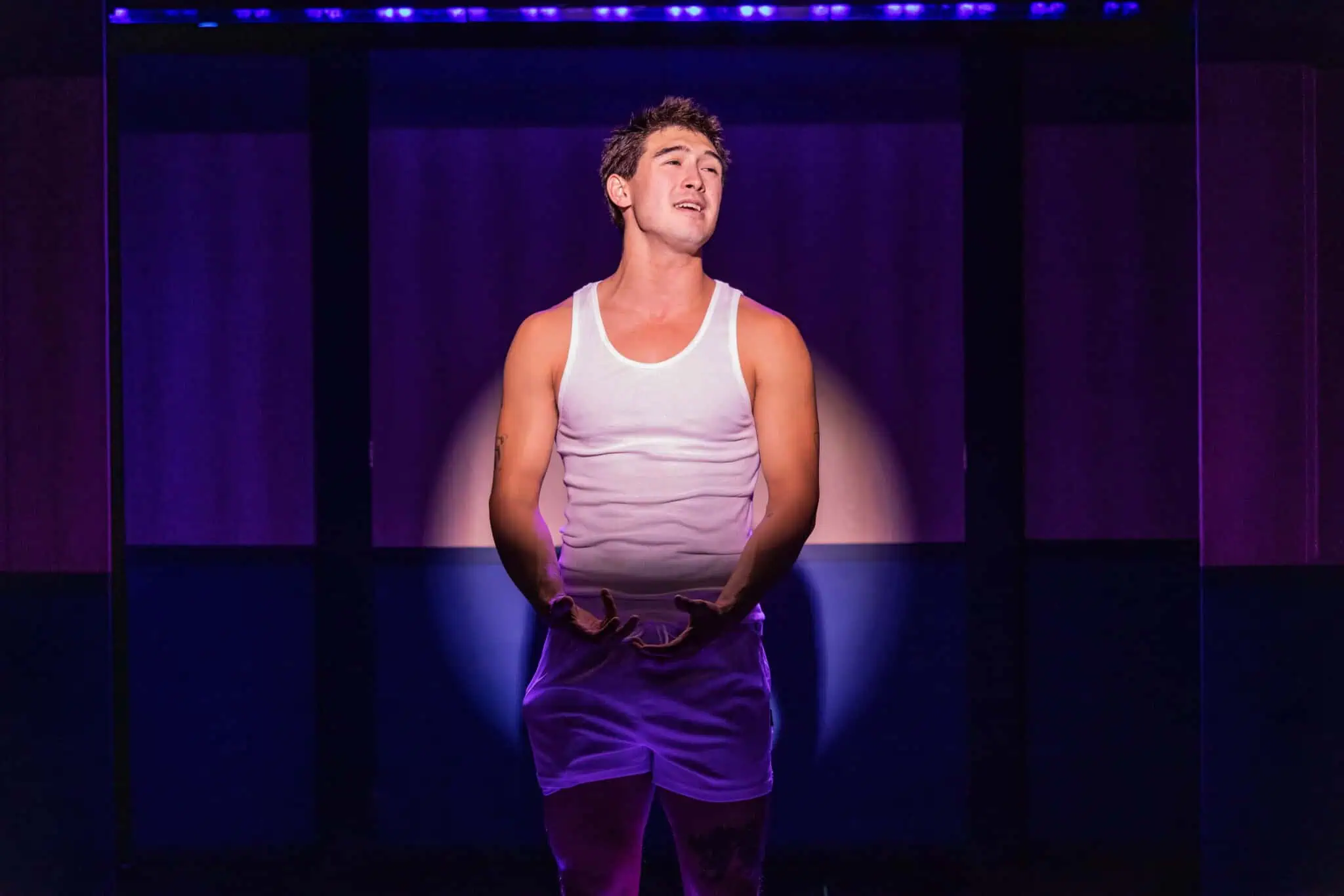 A young man in a white tank top and shorts stands alone on stage under a spotlight, his hands open and expressive as he performs, with a dark background illuminated by purple lighting.