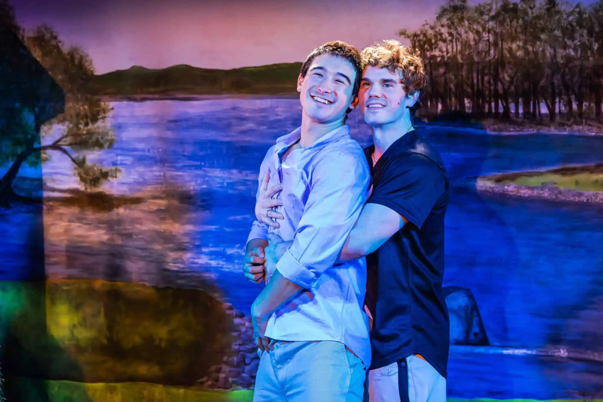 Two young men stand close together, smiling and embracing in front of a brightly painted scenic backdrop of a lake and trees, with one standing behind the other and wrapping his arms around him.