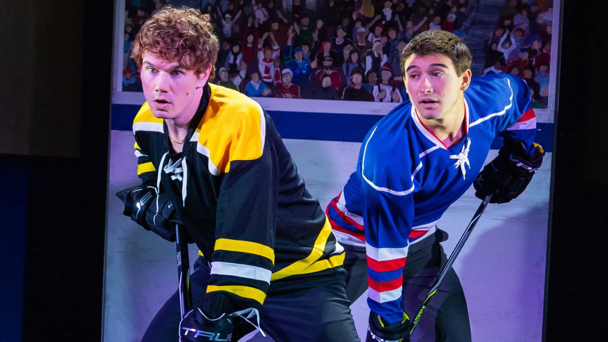 Two hockey players in black/yellow and blue/red uniforms face off on the ice, gripping their sticks and looking intently ahead, with a blurred crowd in the background.