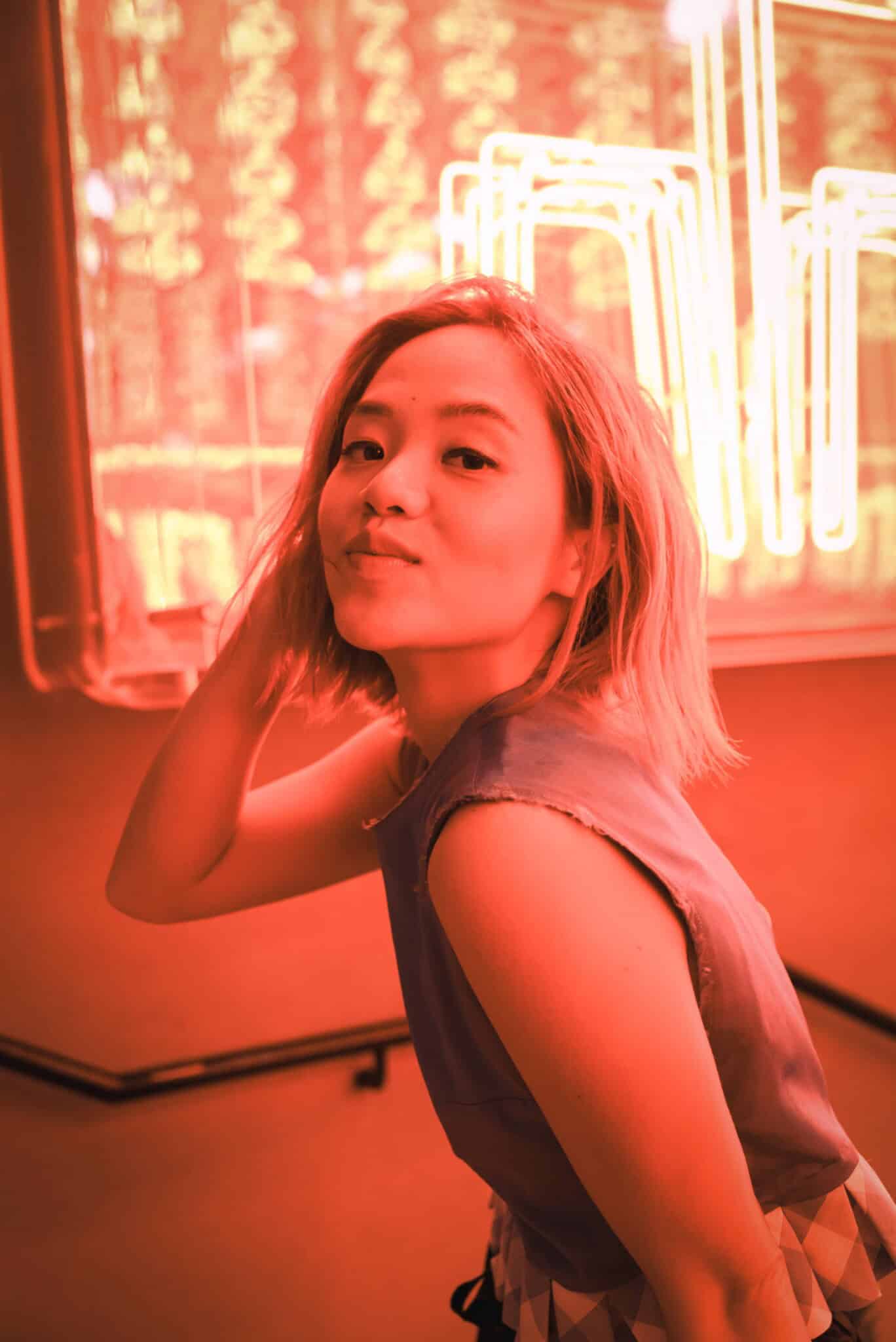A woman with shoulder-length hair poses confidently in front of a bright neon sign, bathed in a vivid orange-red light. She tilts her head and touches her hair, giving a slight smile to the camera.