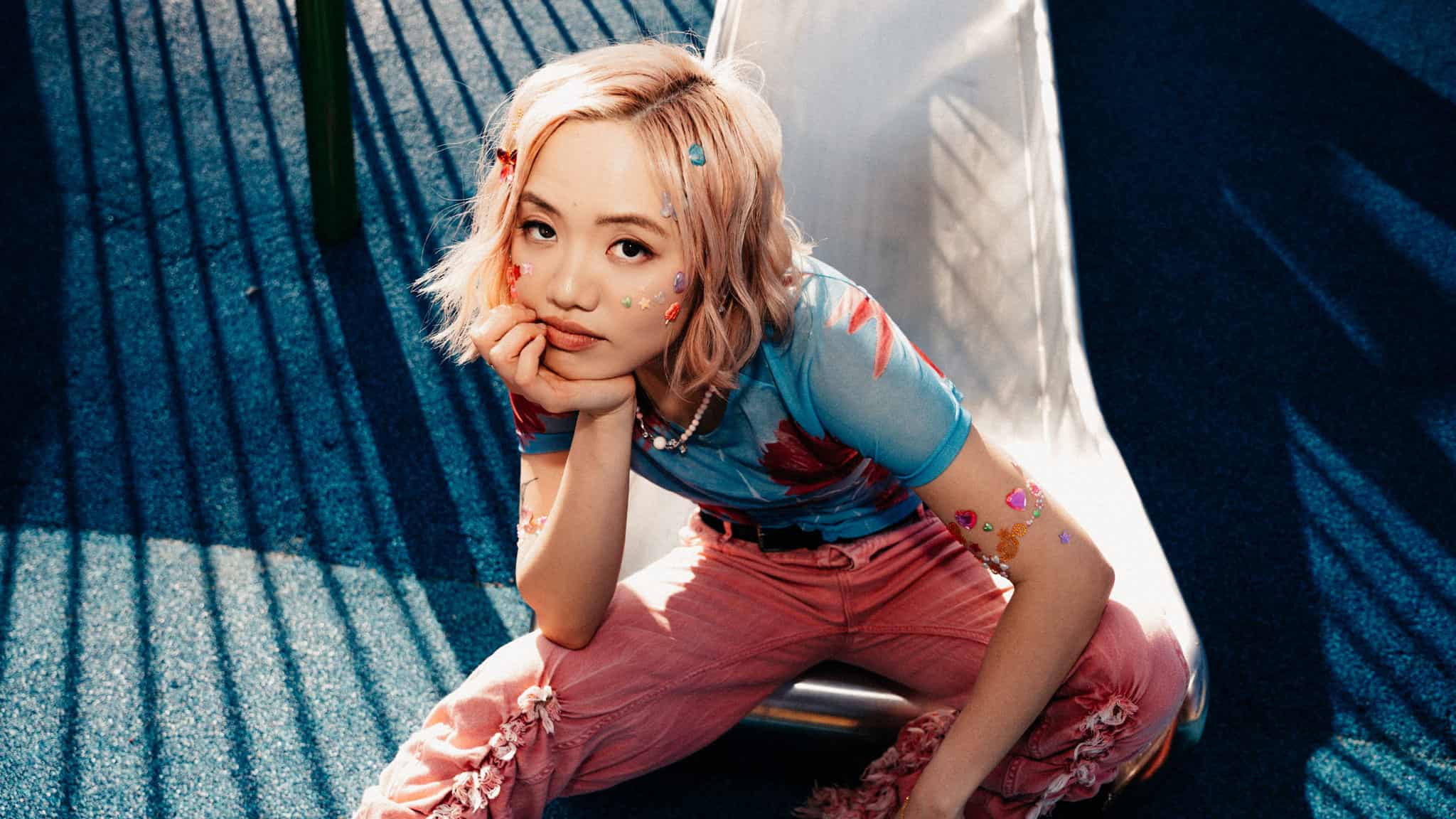 A person with blonde hair sits on a slide, resting their chin on their hand. They wear a blue shirt, pink pants, and colorful stickers on their face and arm, with a thoughtful expression in bright, striped sunlight.