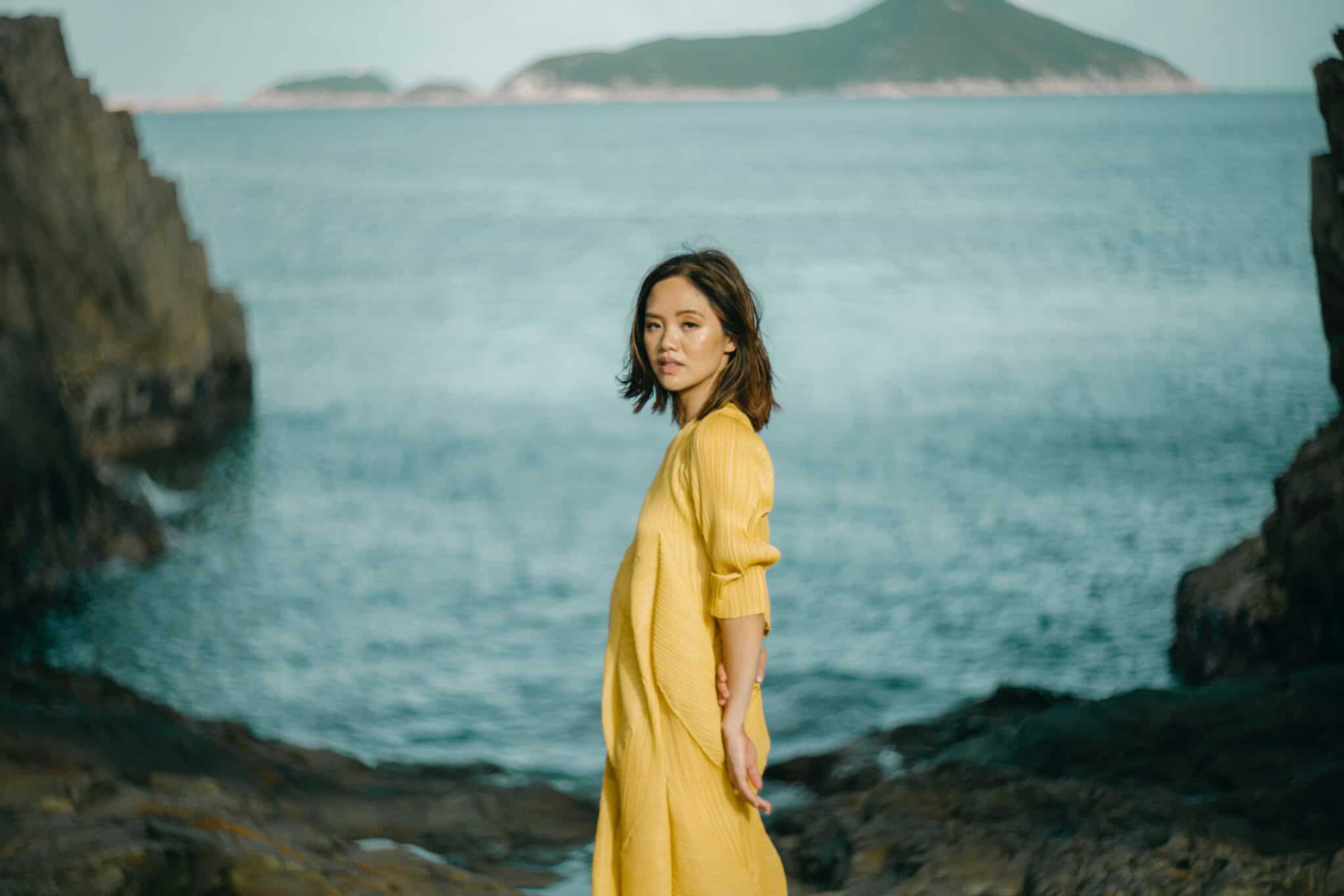 A woman in a yellow dress stands on rocky terrain by the sea, with cliffs on either side and distant islands visible across the water under a partly cloudy sky.