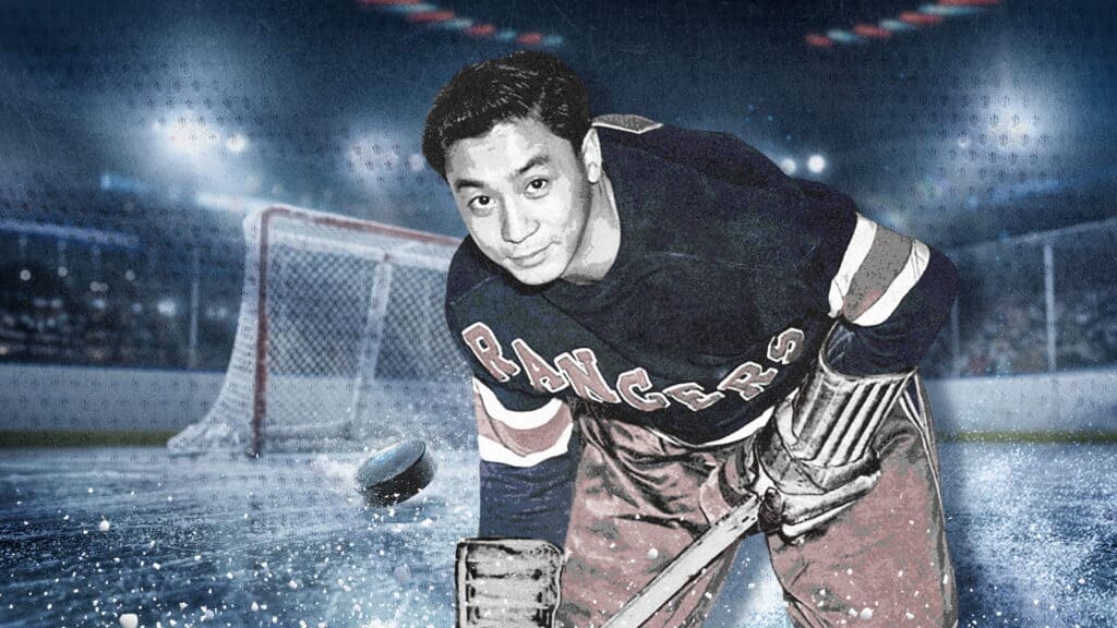 A hockey player wearing a Rangers jersey stands on the ice in front of a goal net, holding a stick and looking at the camera, with a puck and ice particles in the foreground.