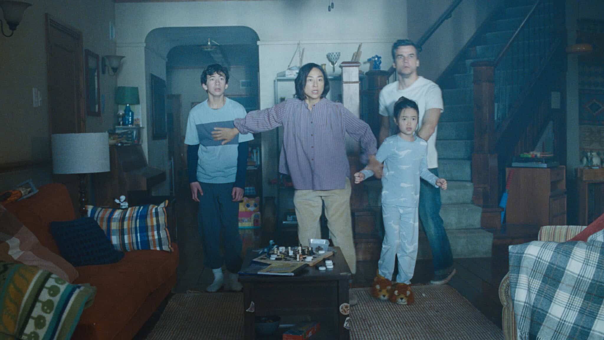 Four people stand tensely in a living room, looking ahead with concern. An adult stands protectively in front of two young people and one man, holding out an arm. The cozy room has a sofa, table, and stairs in the background.