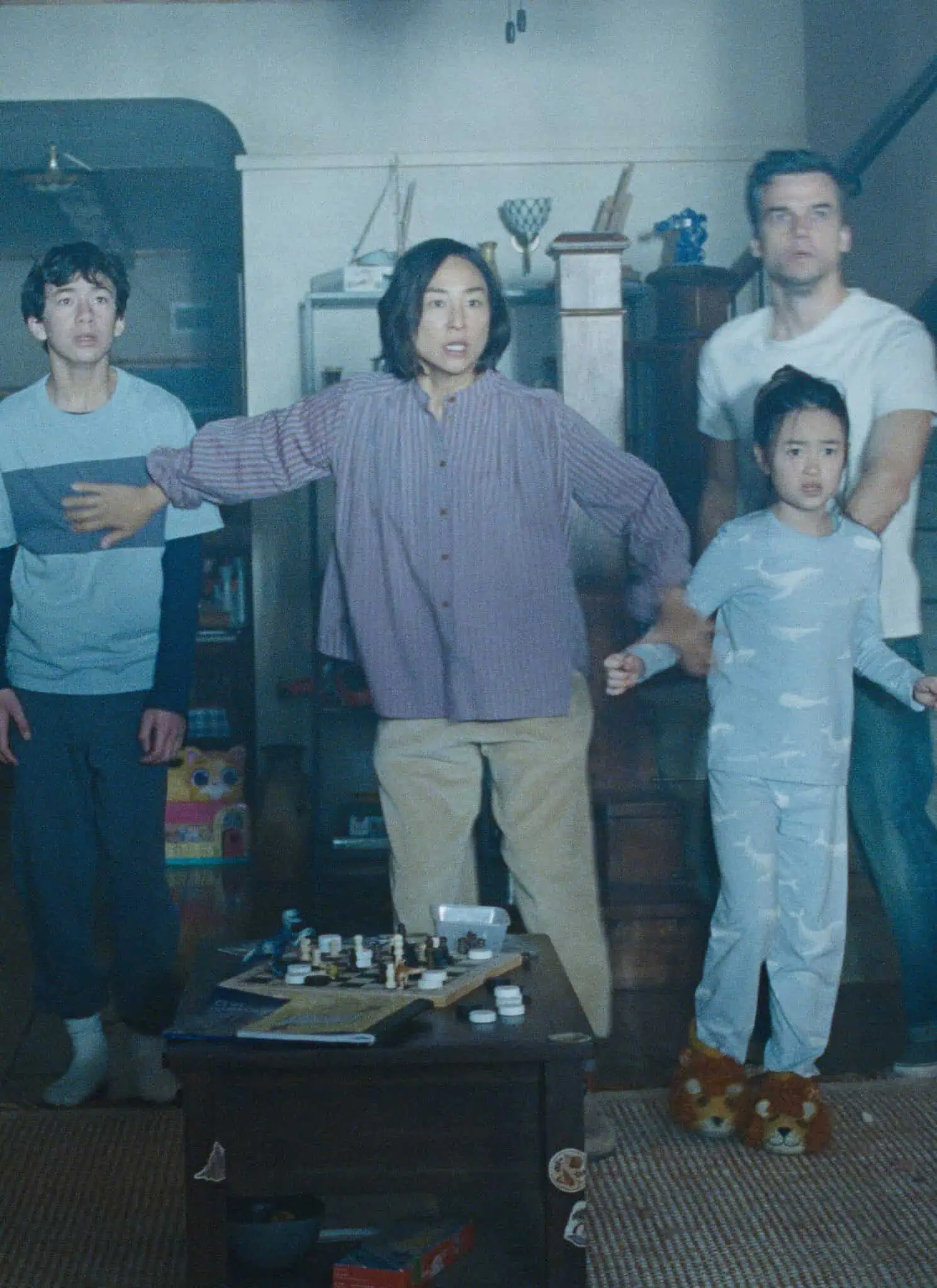 Four people stand tensely in a living room, looking ahead with concern. An adult stands protectively in front of two young people and one man, holding out an arm. The cozy room has a sofa, table, and stairs in the background.