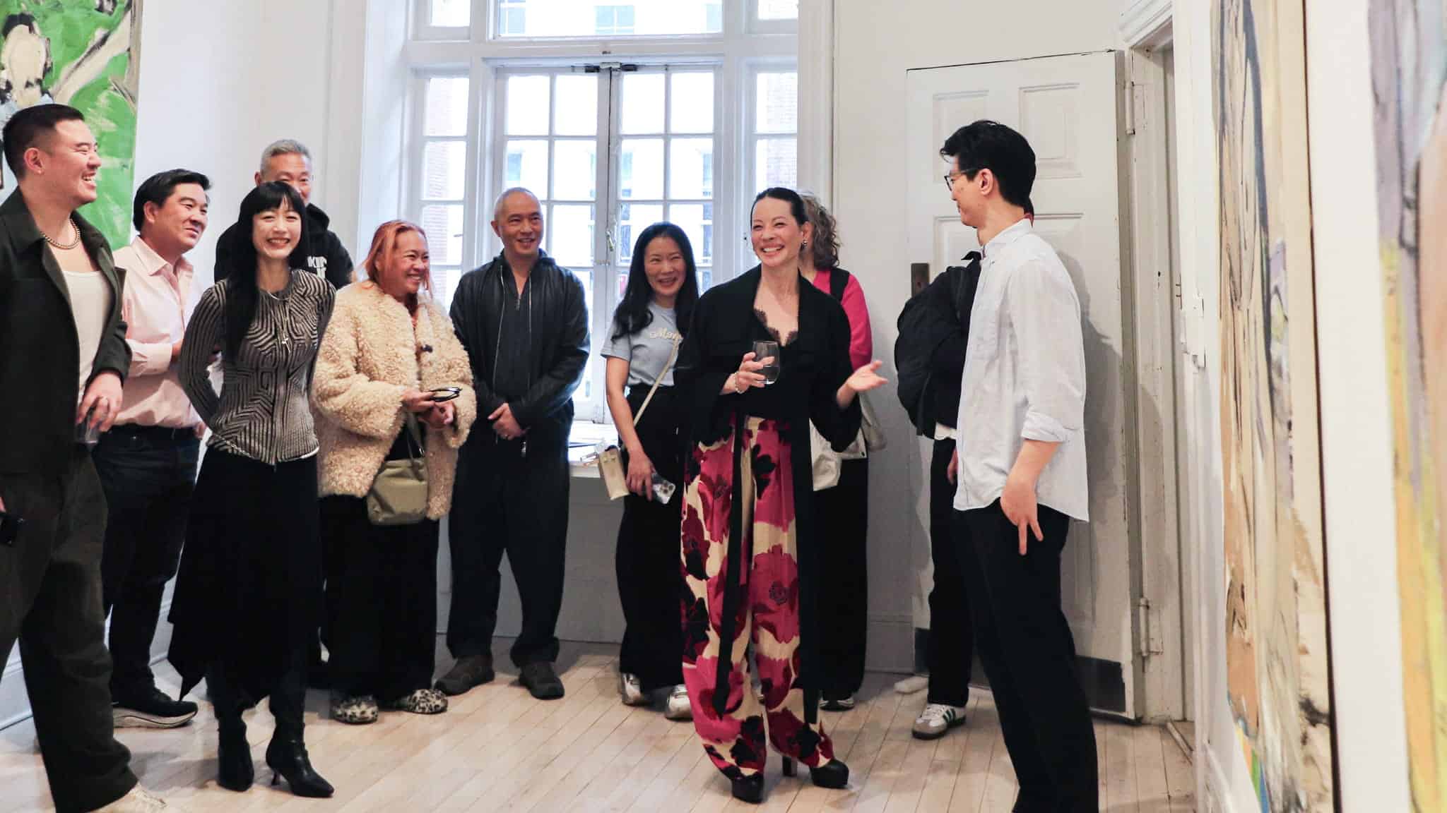 A group of people stand smiling and talking inside an art gallery. One woman in patterned pants gestures while speaking to a man, as others watch and listen, surrounded by paintings and large windows.
