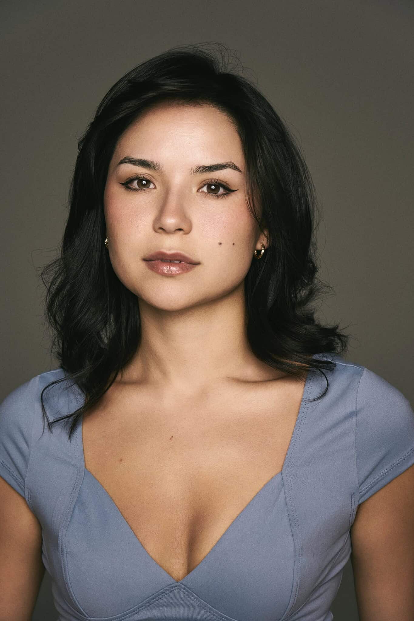 A woman with medium-length dark hair, wearing a light blue V-neck top, looks directly at the camera against a plain, neutral background.
