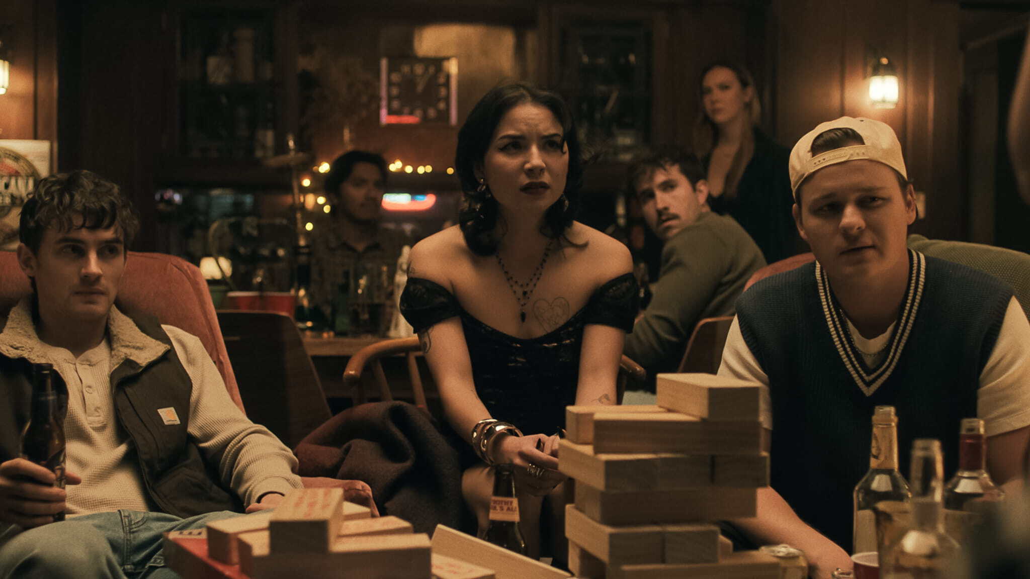 A group of young adults sits around a table stacked with wooden blocks, looking tense and focused. The setting is a dimly lit, cozy room with wooden walls and string lights in the background.