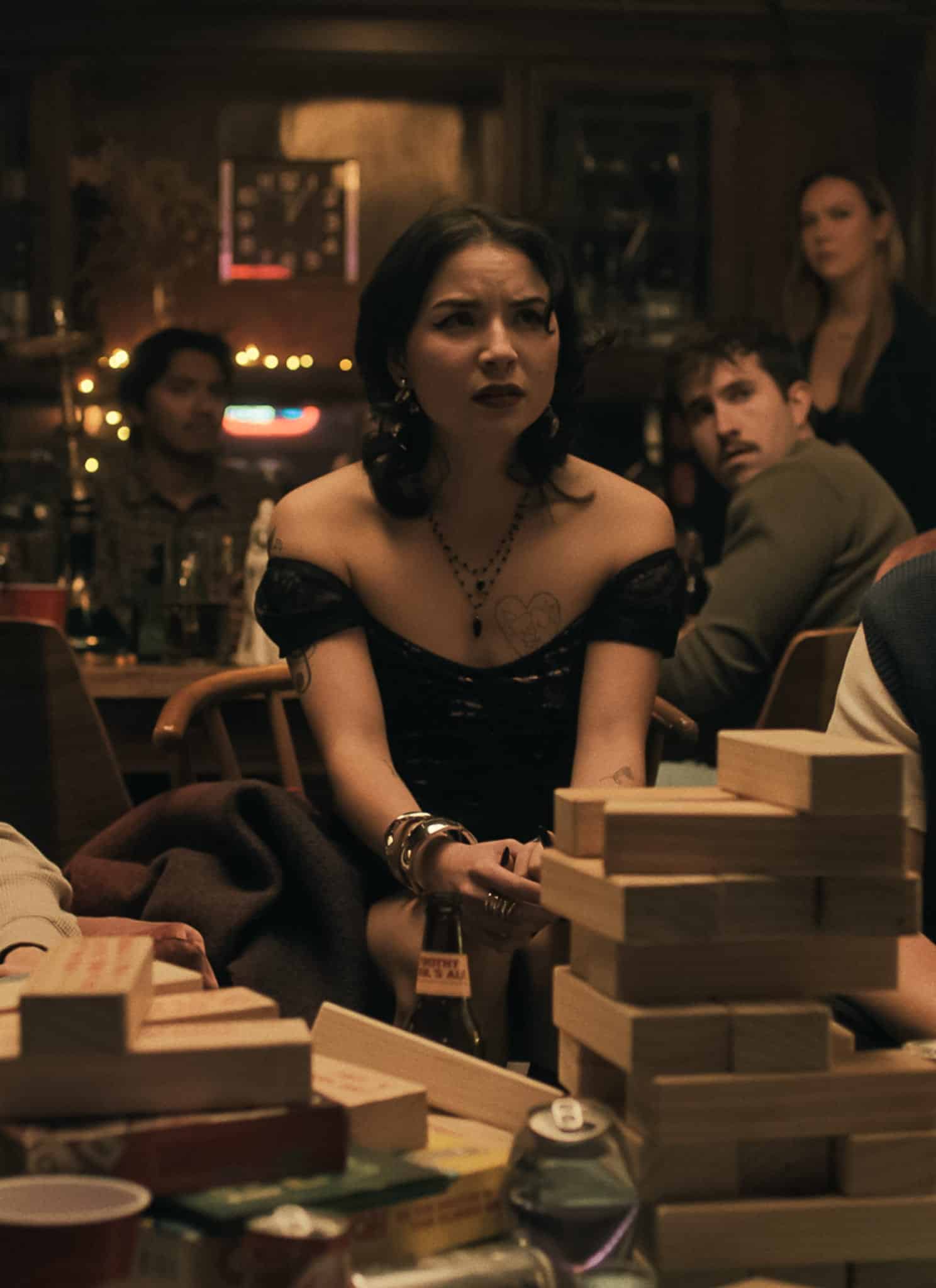 A group of young adults sits around a table stacked with wooden blocks, looking tense and focused. The setting is a dimly lit, cozy room with wooden walls and string lights in the background.