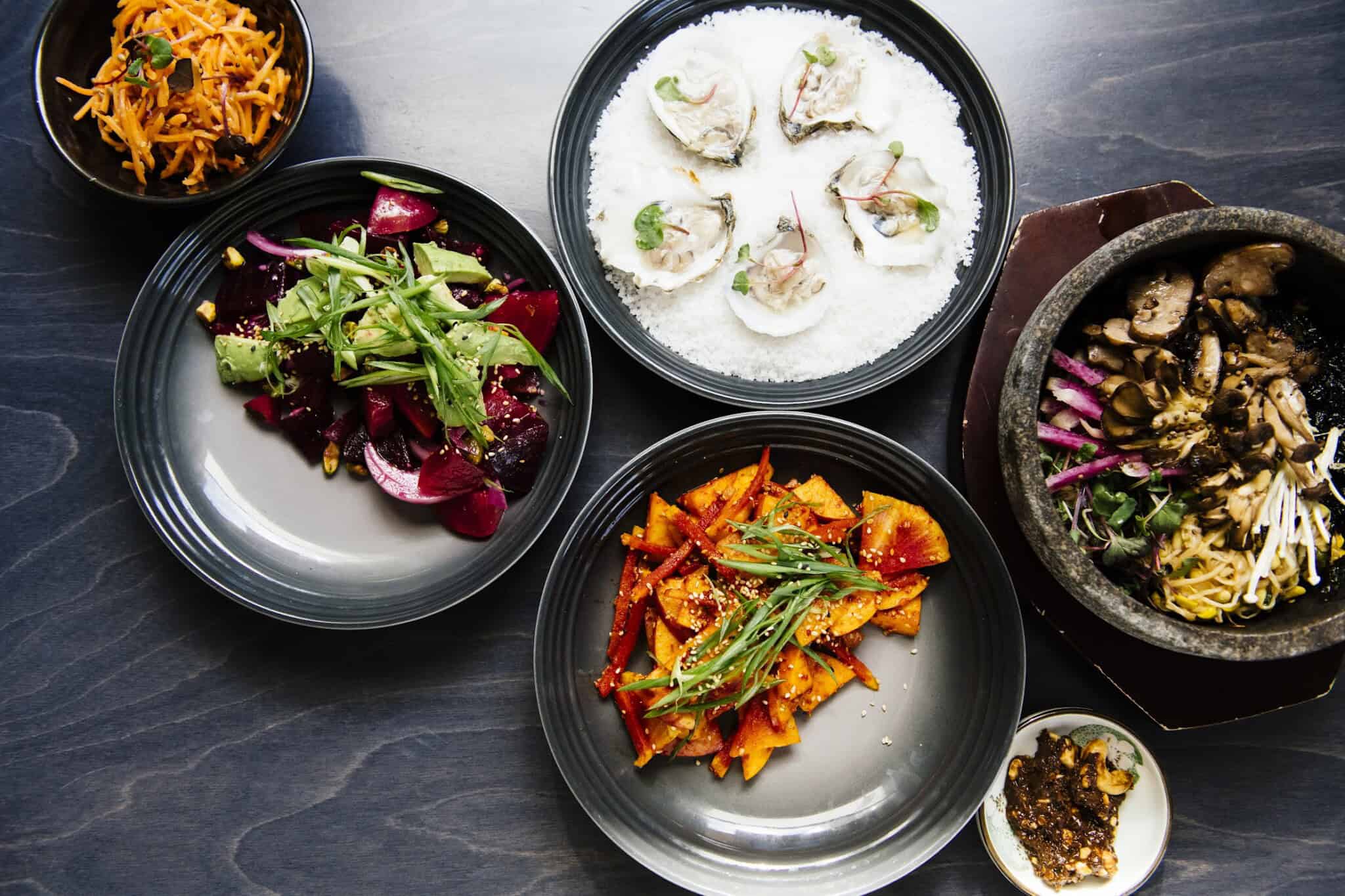 An overhead view of assorted dishes: oysters on ice, mixed greens with beets, spicy carrot salad, a vegetable and meat rice bowl, chili sauce, and a plate of orange vegetables on a dark tabletop.