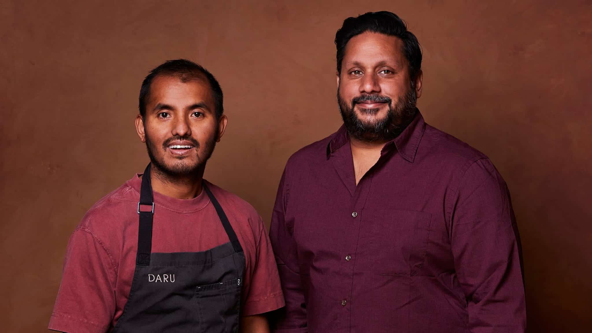Two men, Suresh Sundas and Dante Datta, stand side by side in front of a brown background. One wears a maroon shirt and dark apron labeled DARU, while the other sports a maroon button-up. Both are smiling and facing the camera.