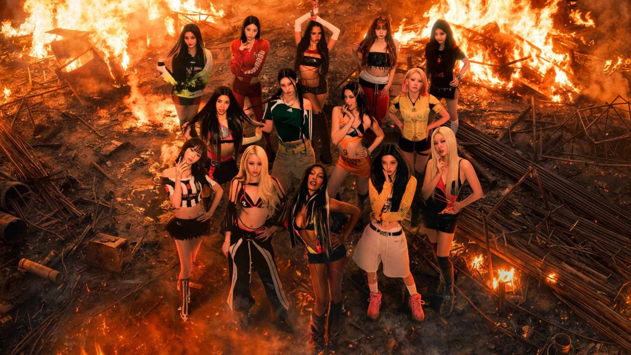 Thirteen women in stylish outfits stand together in a circle, looking up at the camera, surrounded by burning debris and intense flames in a dramatic, fiery setting.