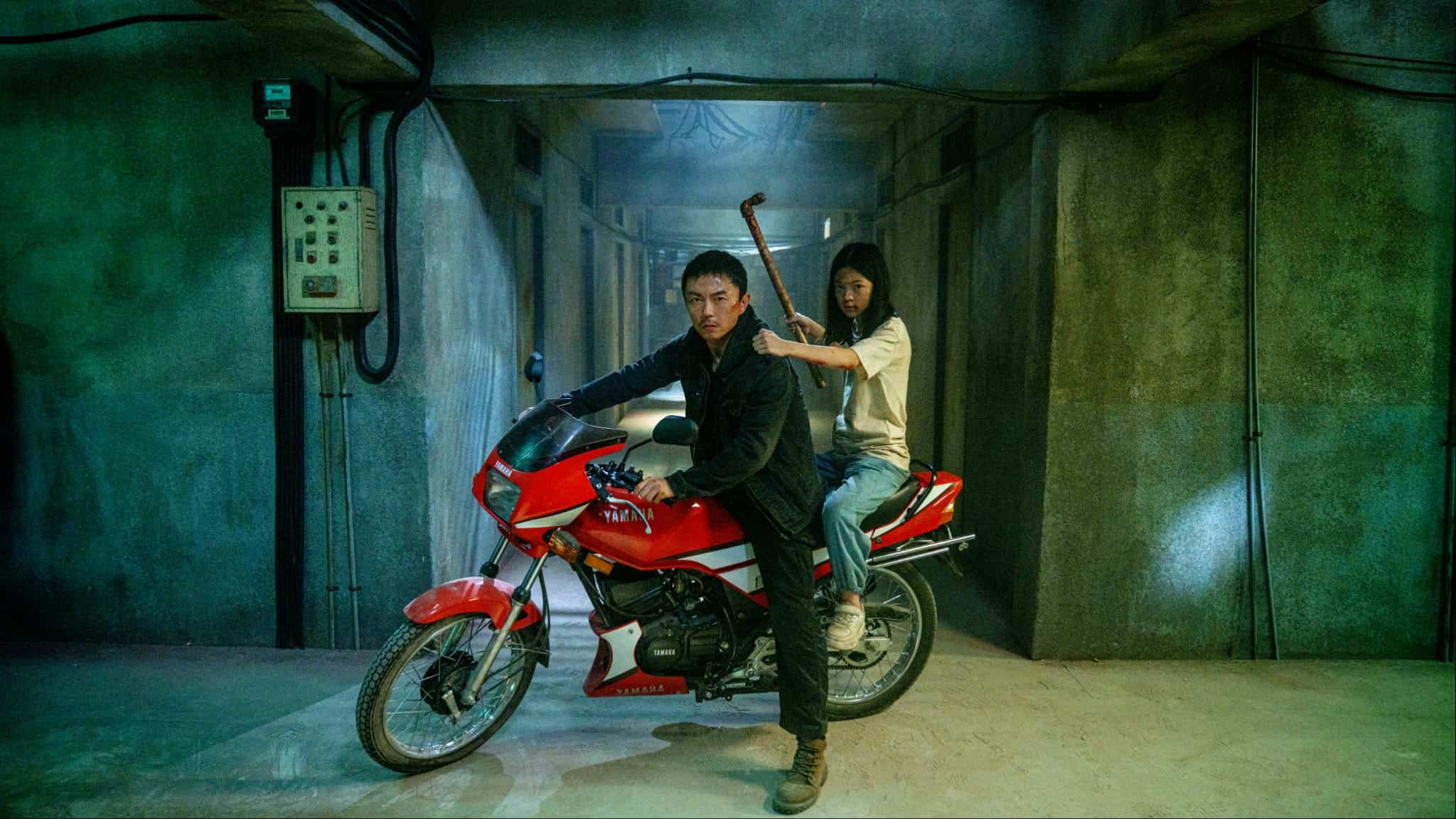 A man sits on a red motorcycle in a dimly lit concrete hallway, while a woman sits behind him holding a crowbar, both looking serious and alert.
