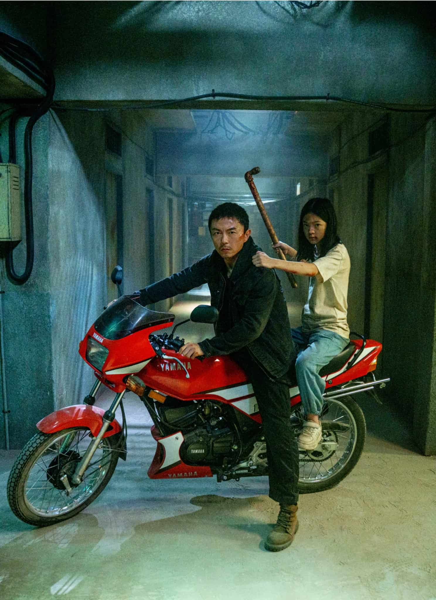 A man sits on a red motorcycle in a dimly lit concrete hallway, while a woman sits behind him holding a crowbar, both looking serious and alert.